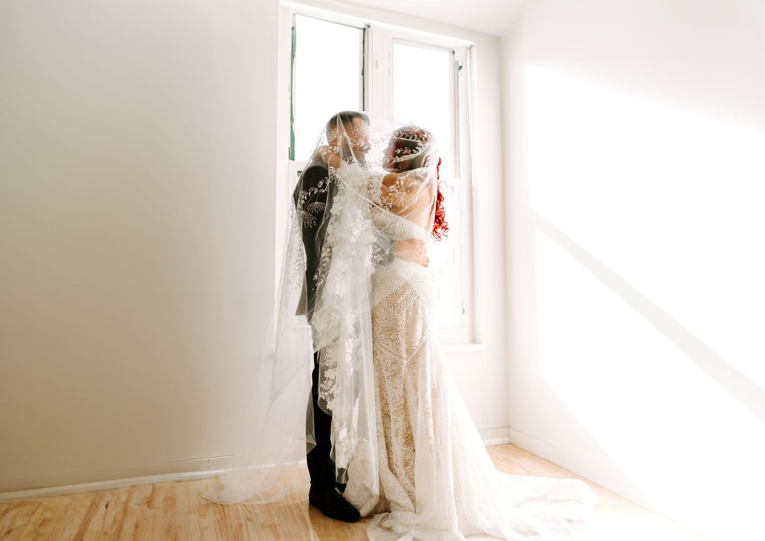 A romantic couple in wedding attire stands near a bright window in an elegant minimalist white room.