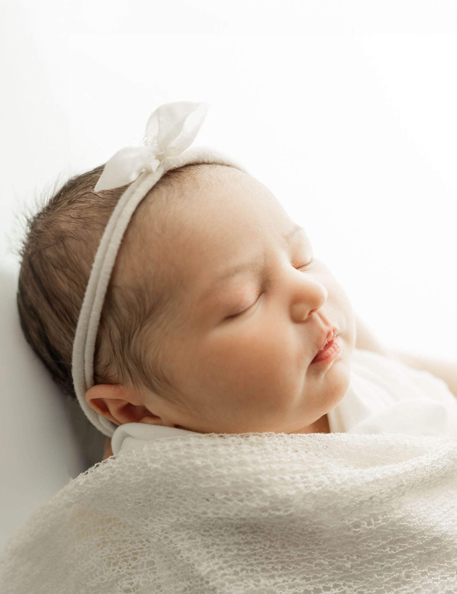 Close-up portrait of a sleeping newborn wearing a white headband and wrapped in a soft blanket.