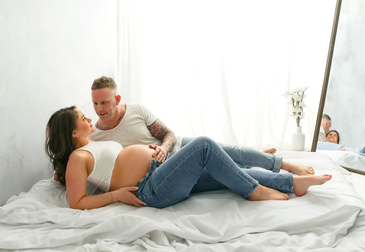A serene maternity portrait featuring a couple relaxing together on a bed in front of a bright window.