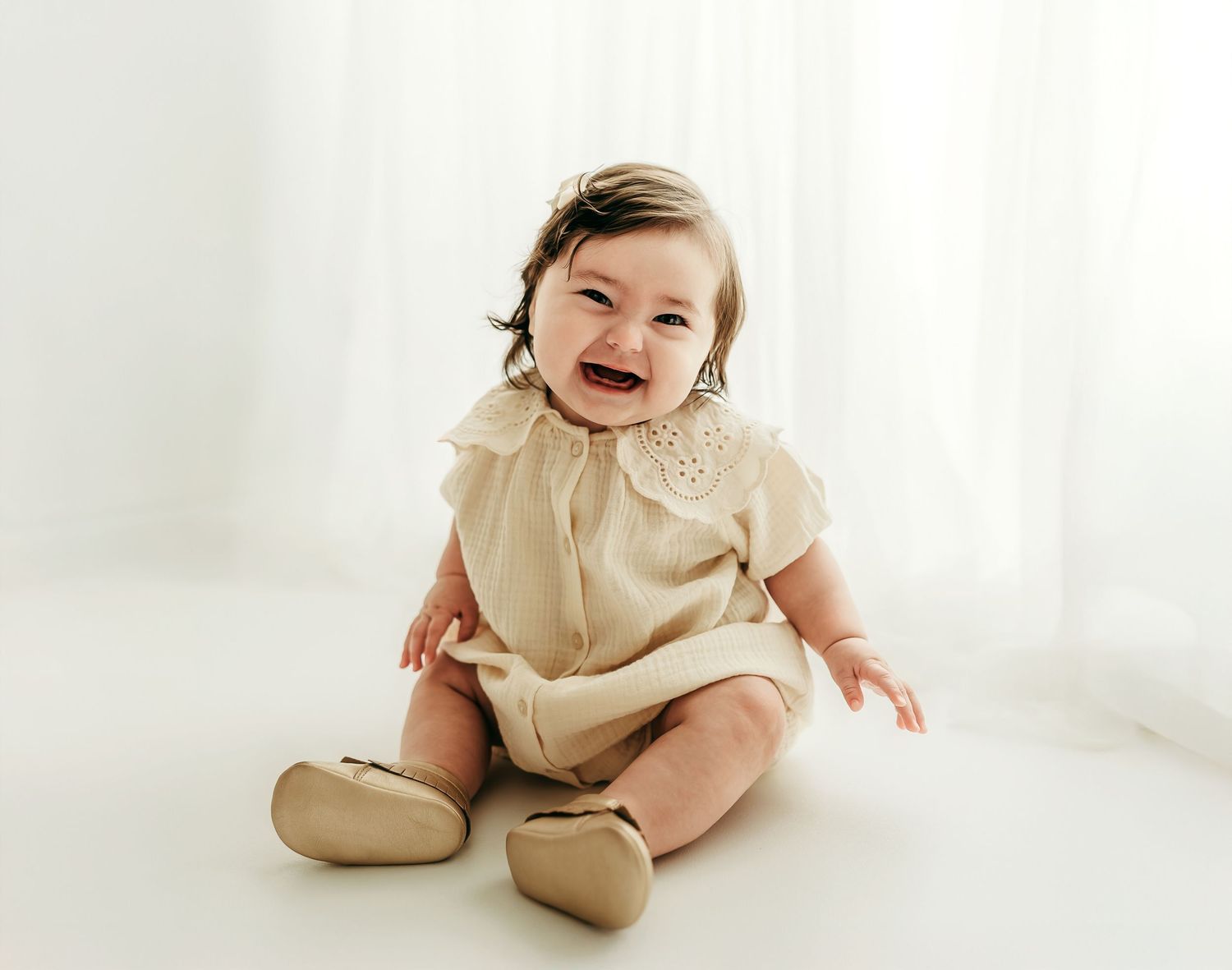 A smiling 6 month old baby girl sitting on a white backdrop during a soft, neutral milestone photo session.