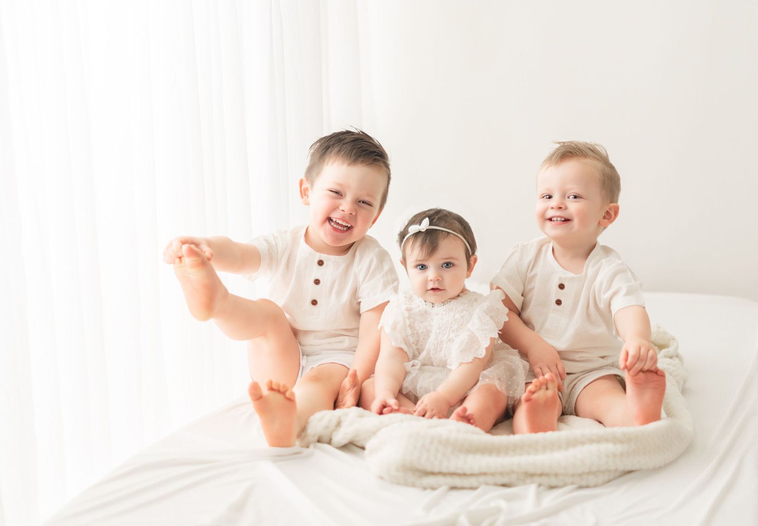 Three babies in white outfits sit together on white bedding while laughing and playing.