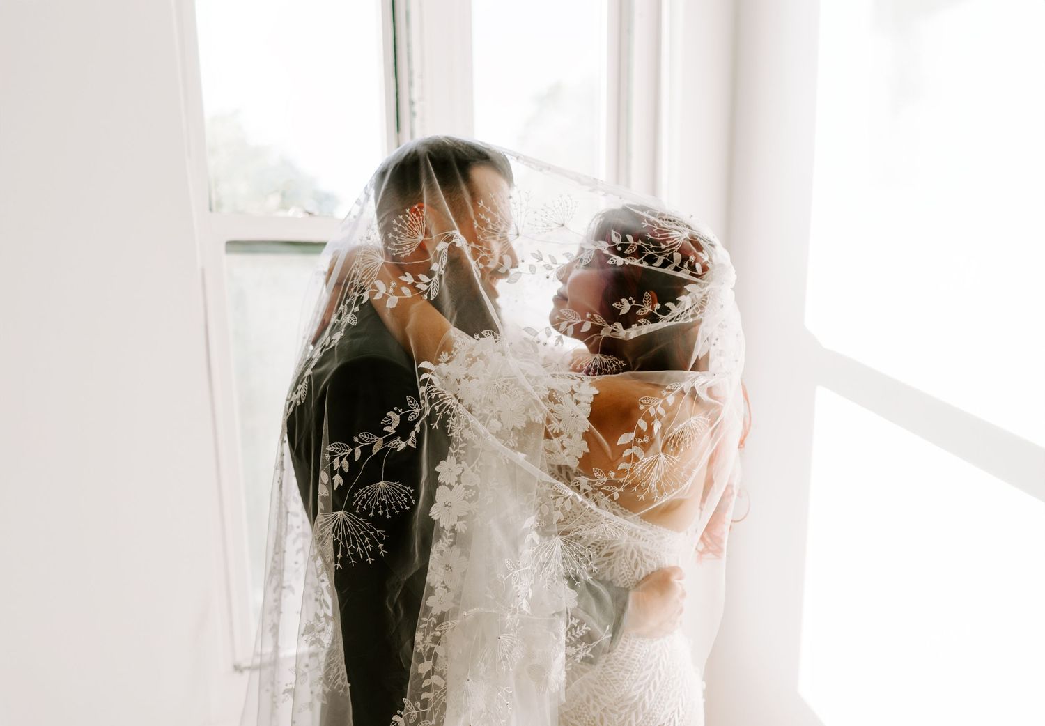 Romantic wedding portrait with couple embracing under delicate lace veils by window.