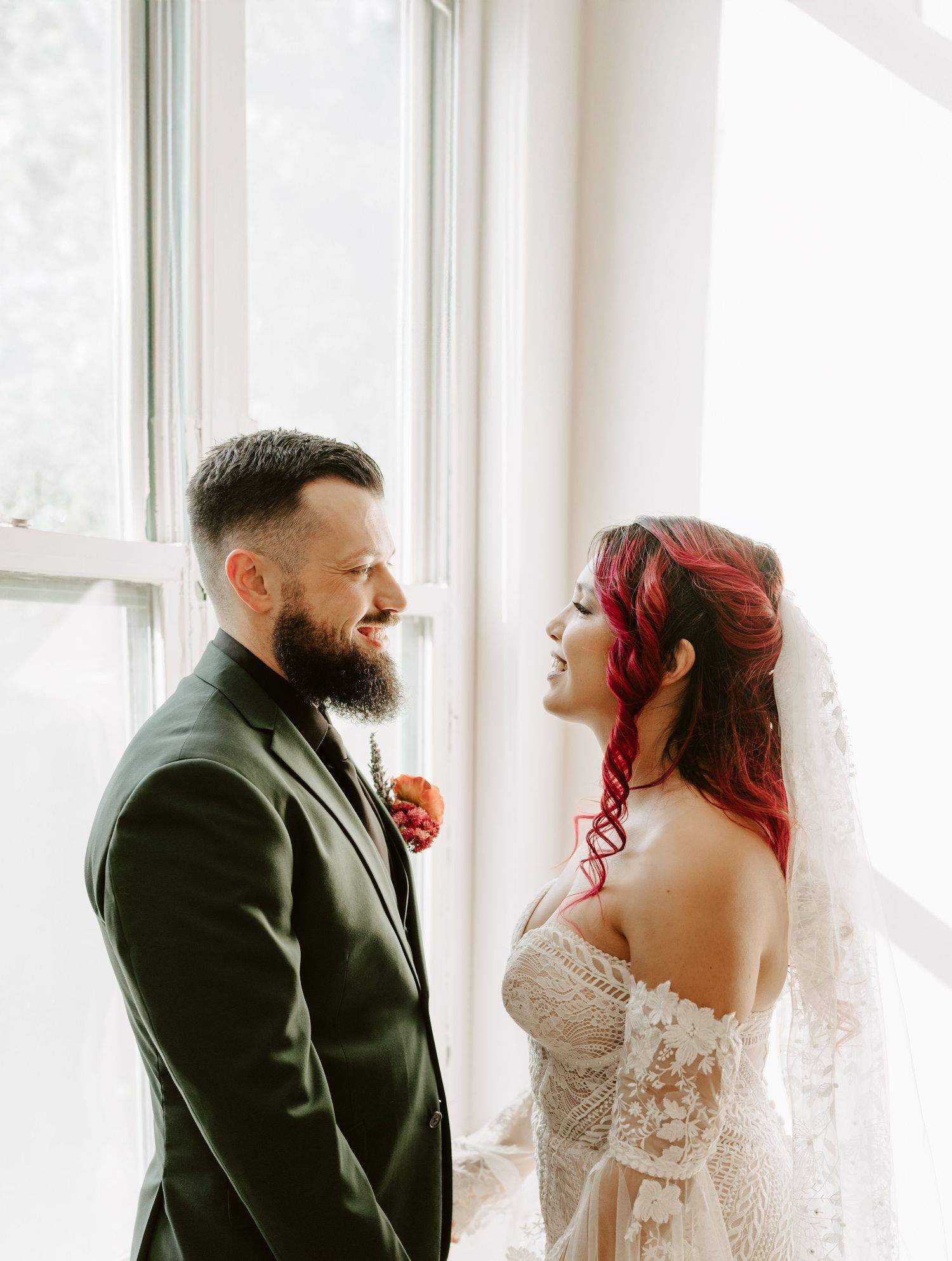 A smiling couple in wedding attire stand next to bright windows, with the bride wearing red hair and a white lace gown.