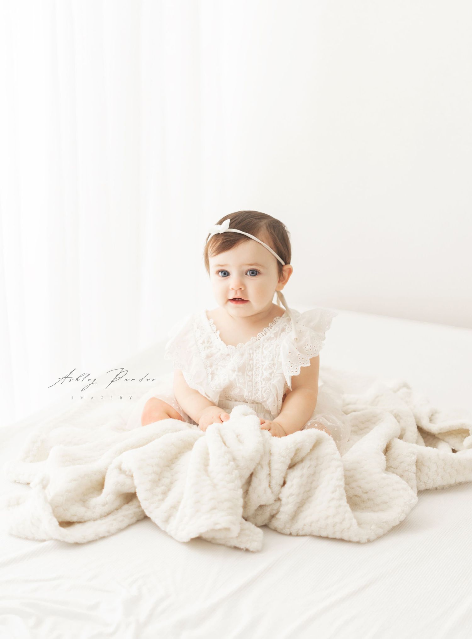 A baby wearing a lace dress sits on white bedding in a bright, minimalist setting for a portrait photo session.