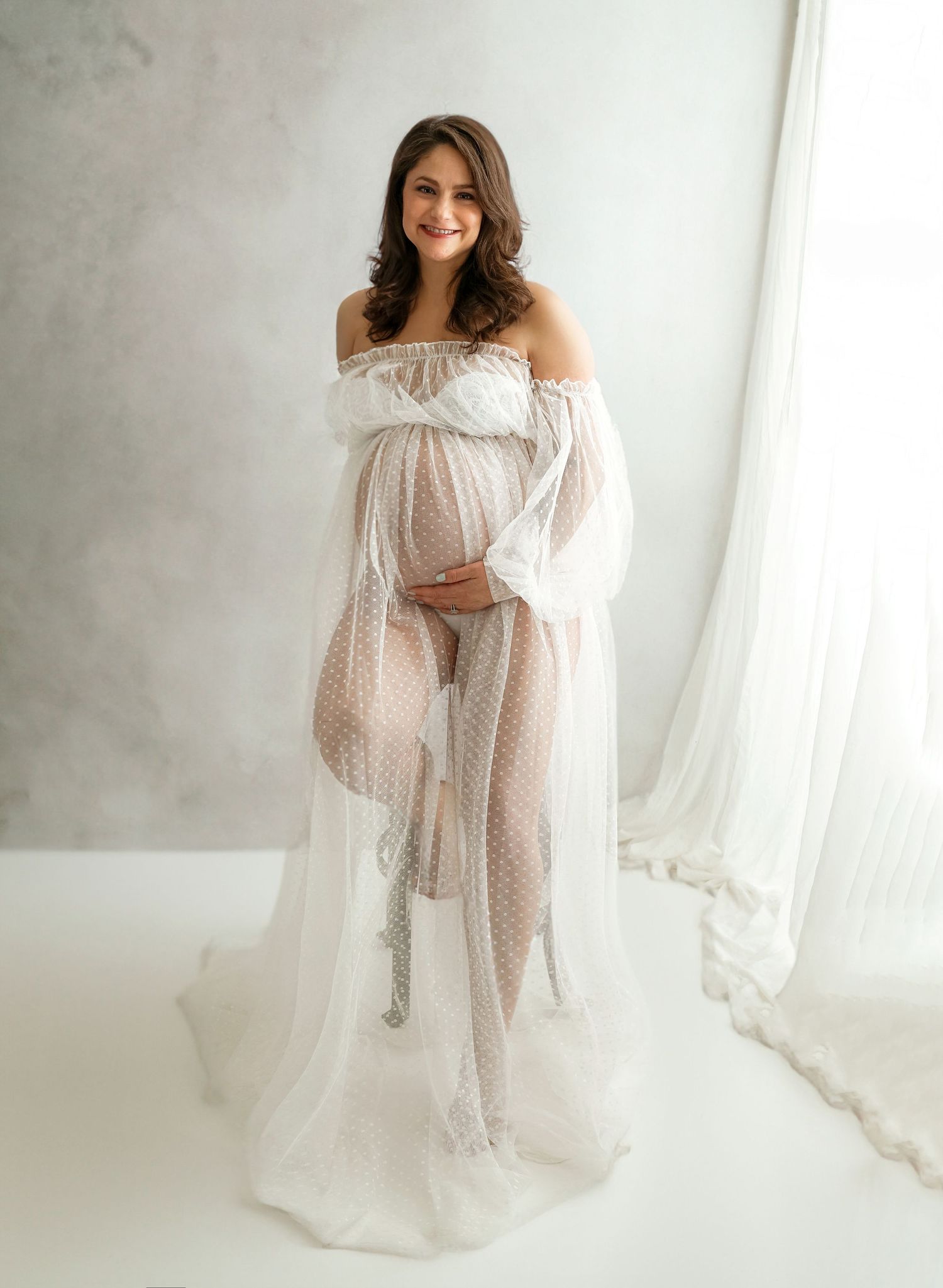 An elegant maternity portrait in a flowing white off-shoulder gown against a light background.
