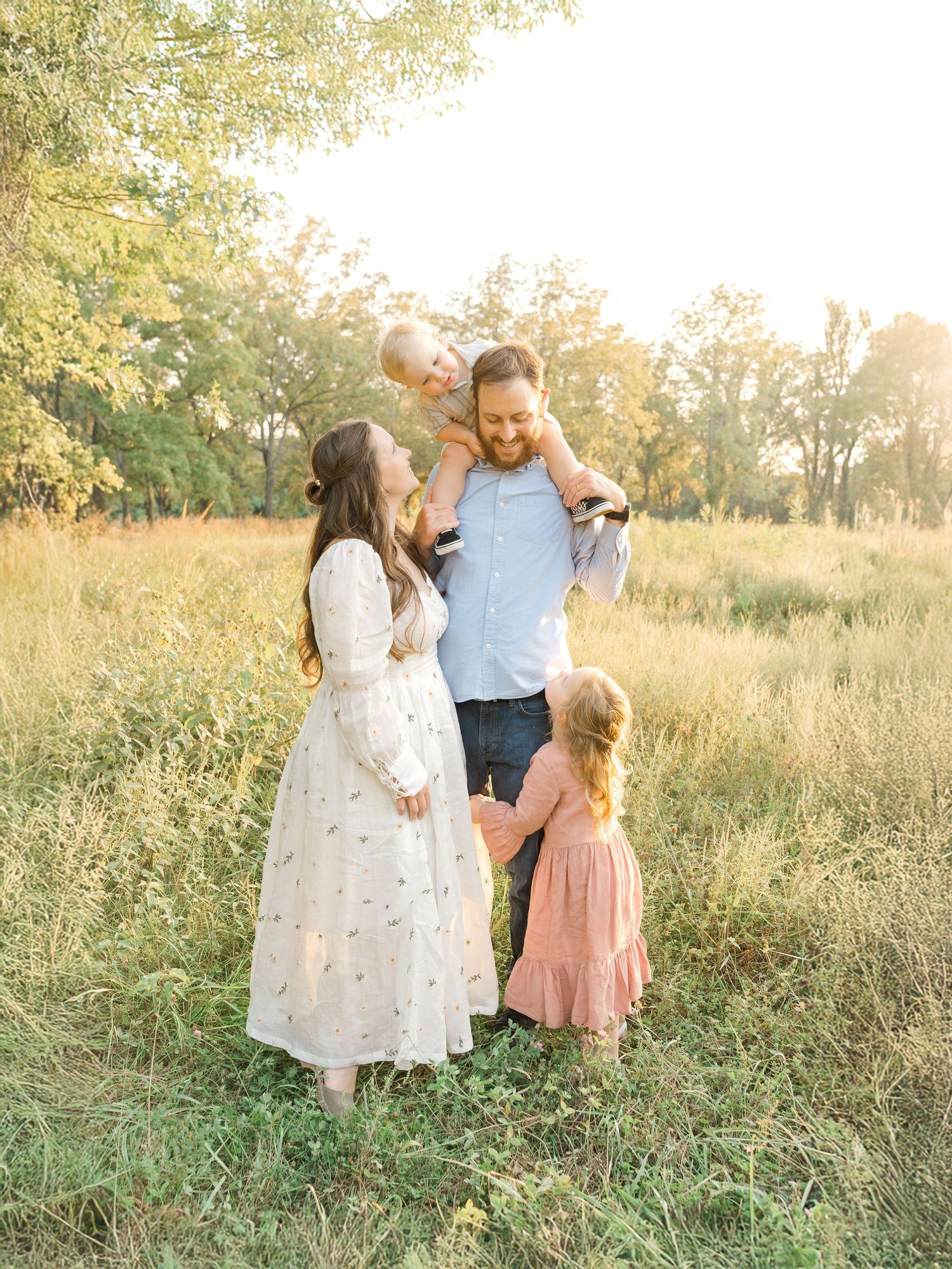 A family of four standing together in a grassy field at sunset, with a father holding a child on his shoulders during an outdoor session in