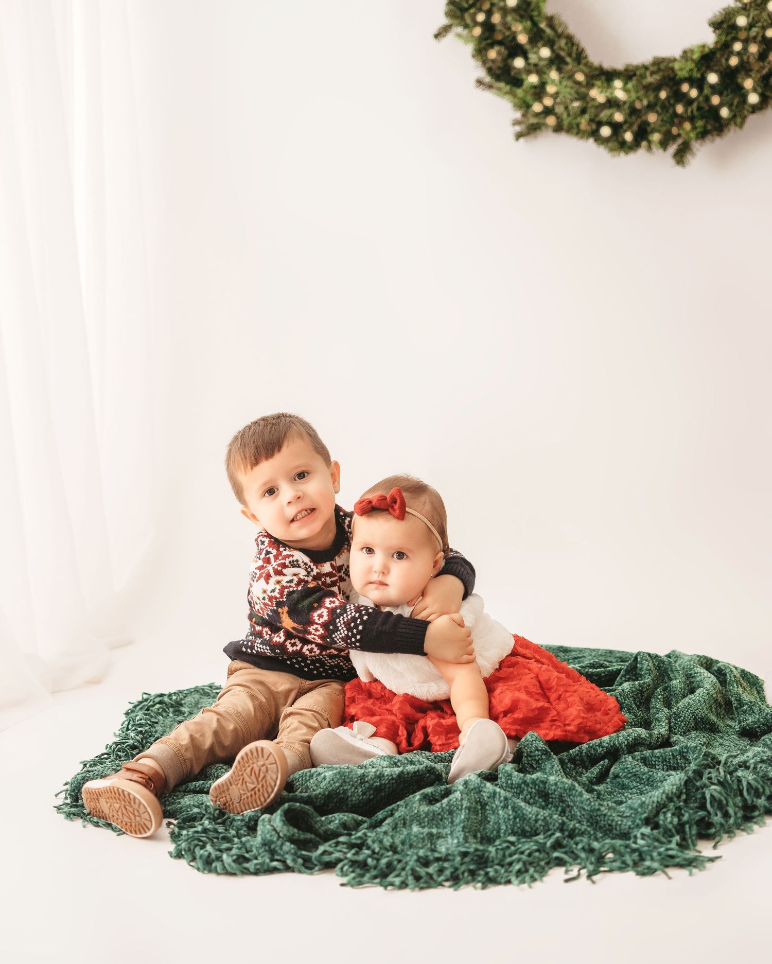 Two children sit together on a green blanket against a white backdrop with a holiday wreath on the wall.