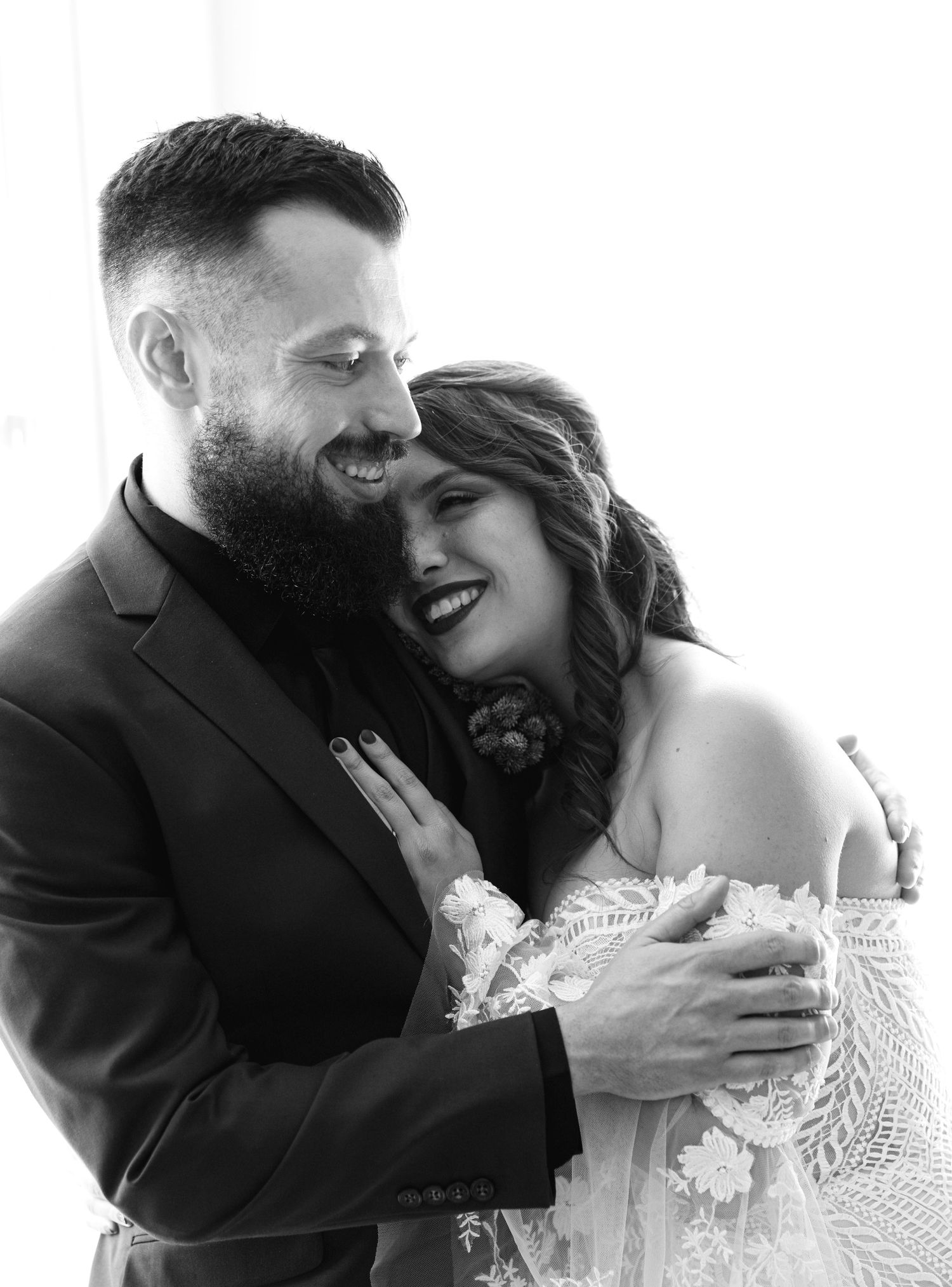 Black and white wedding portrait capturing an intimate moment between a bride in lace and groom in suit sharing laughter.