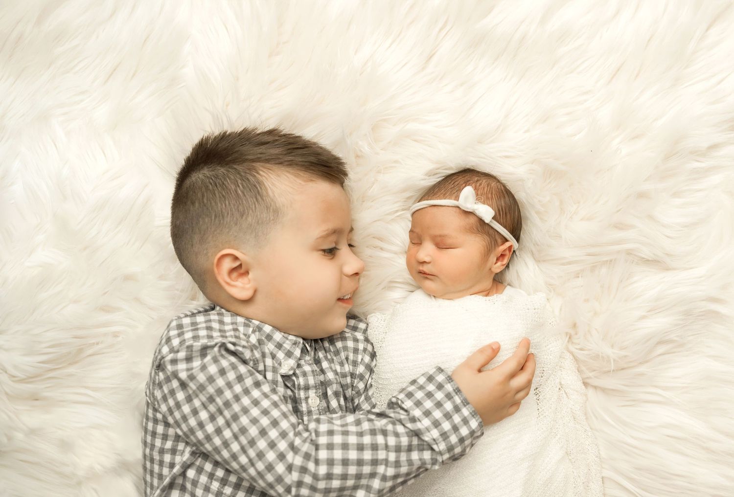 Two siblings lay on white fur, an older child in plaid shirt next to a sleeping newborn wearing a white headband.