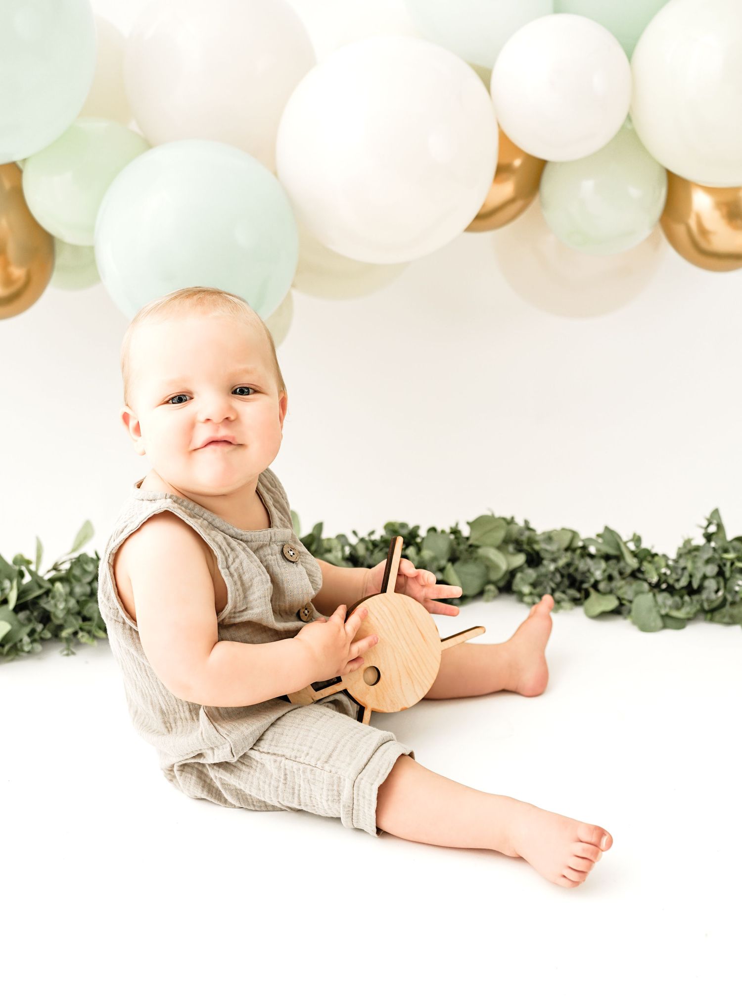A baby in a linen romper sits on white backdrop with mint and gold balloons and eucalyptus garland decorations.