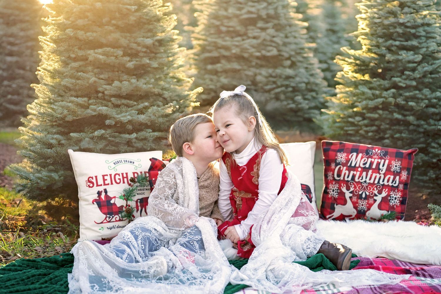 A young boy kisses his sister while sitting on a festive blanket with Christmas-themed pillows