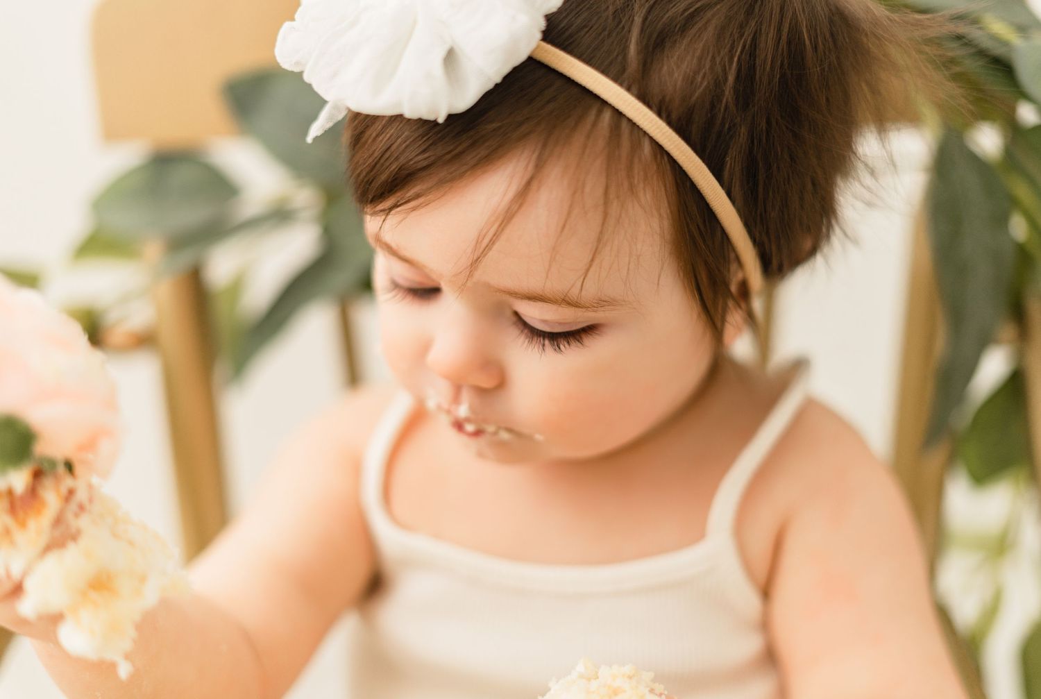 A small child in a white dress and headband looks down while surrounded by soft greenery and warm lighting.
