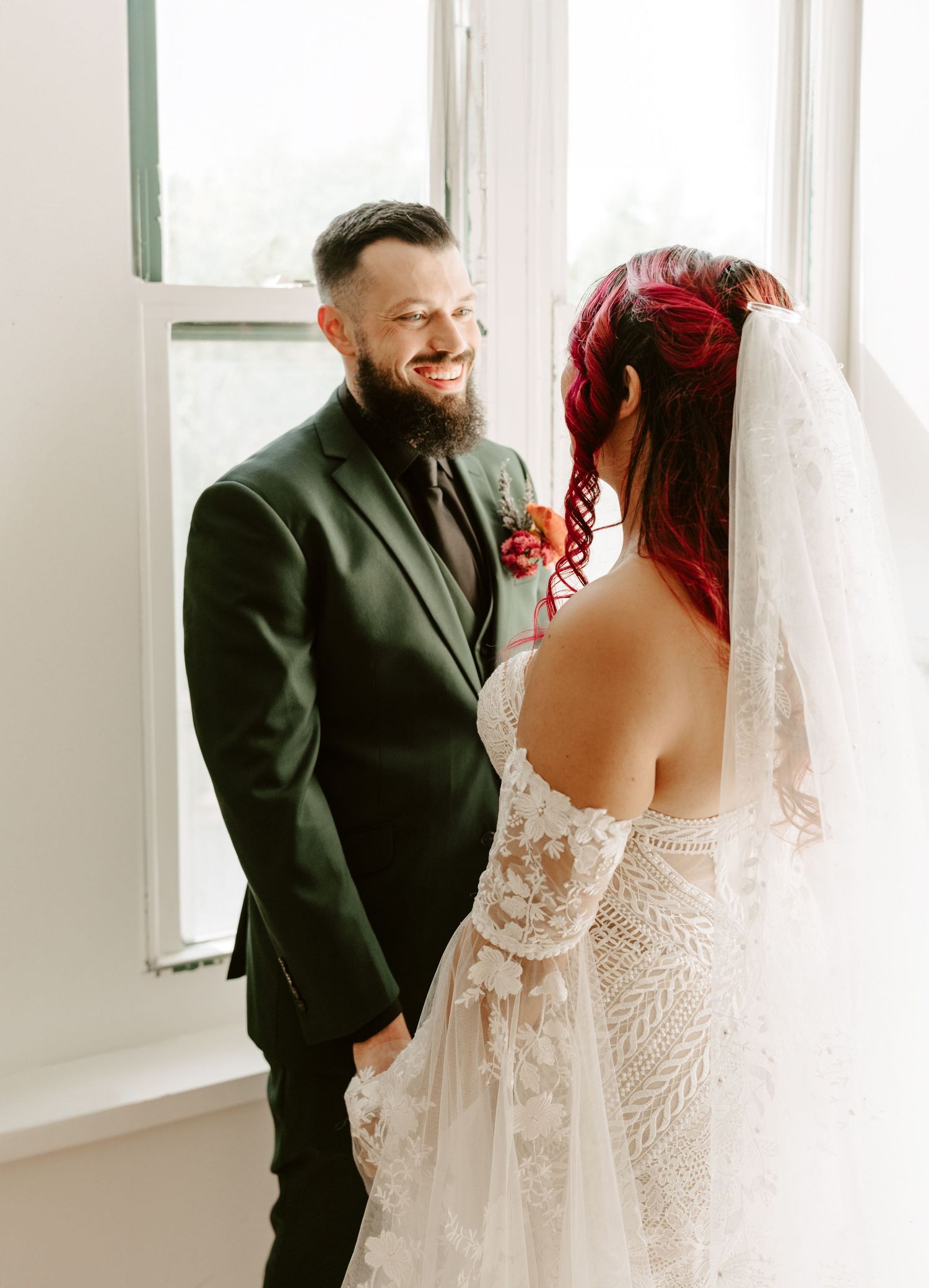 A couple shares an intimate moment in front of a window, with the bride in a lace dress and groom in dark suit.