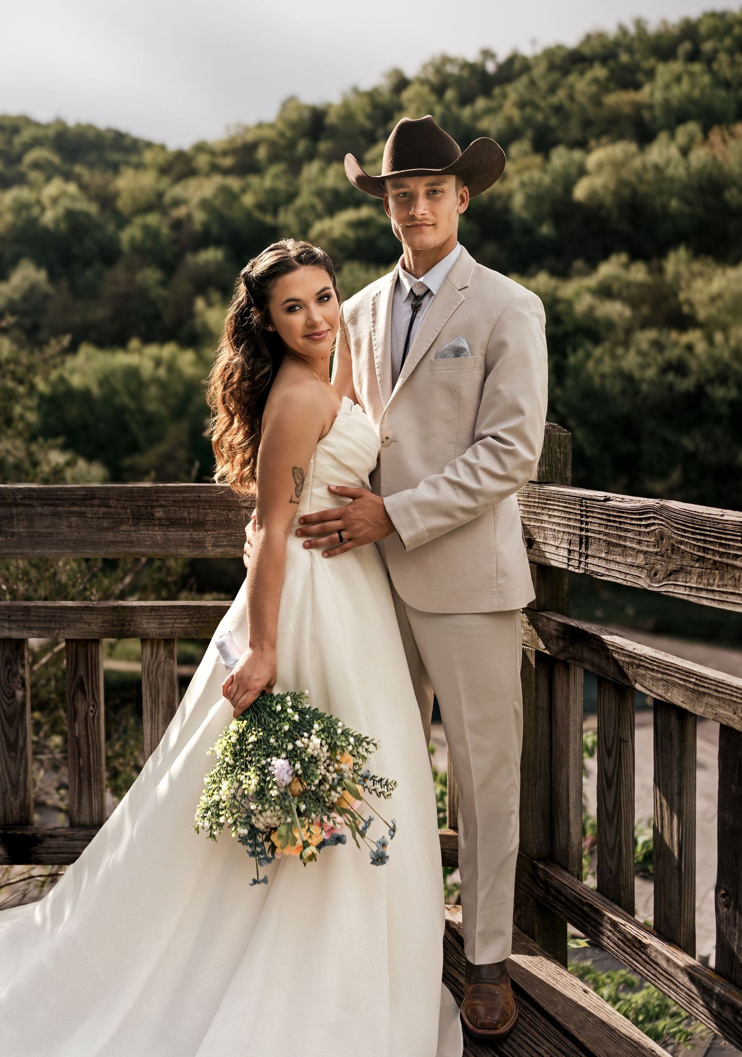 A bride in a white gown holding a wildflower bouquet poses with a groom in a tan suit and cowboy hat on a wooden bridge.
