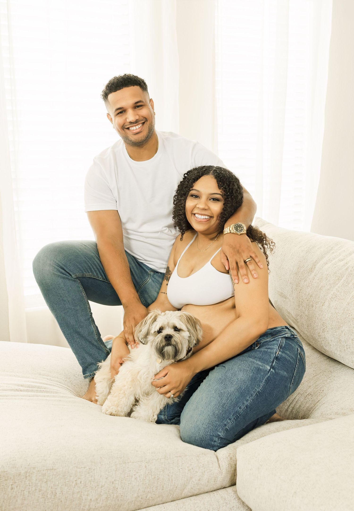 A couple relaxes on a white couch with their small dog in a bright, airy room.