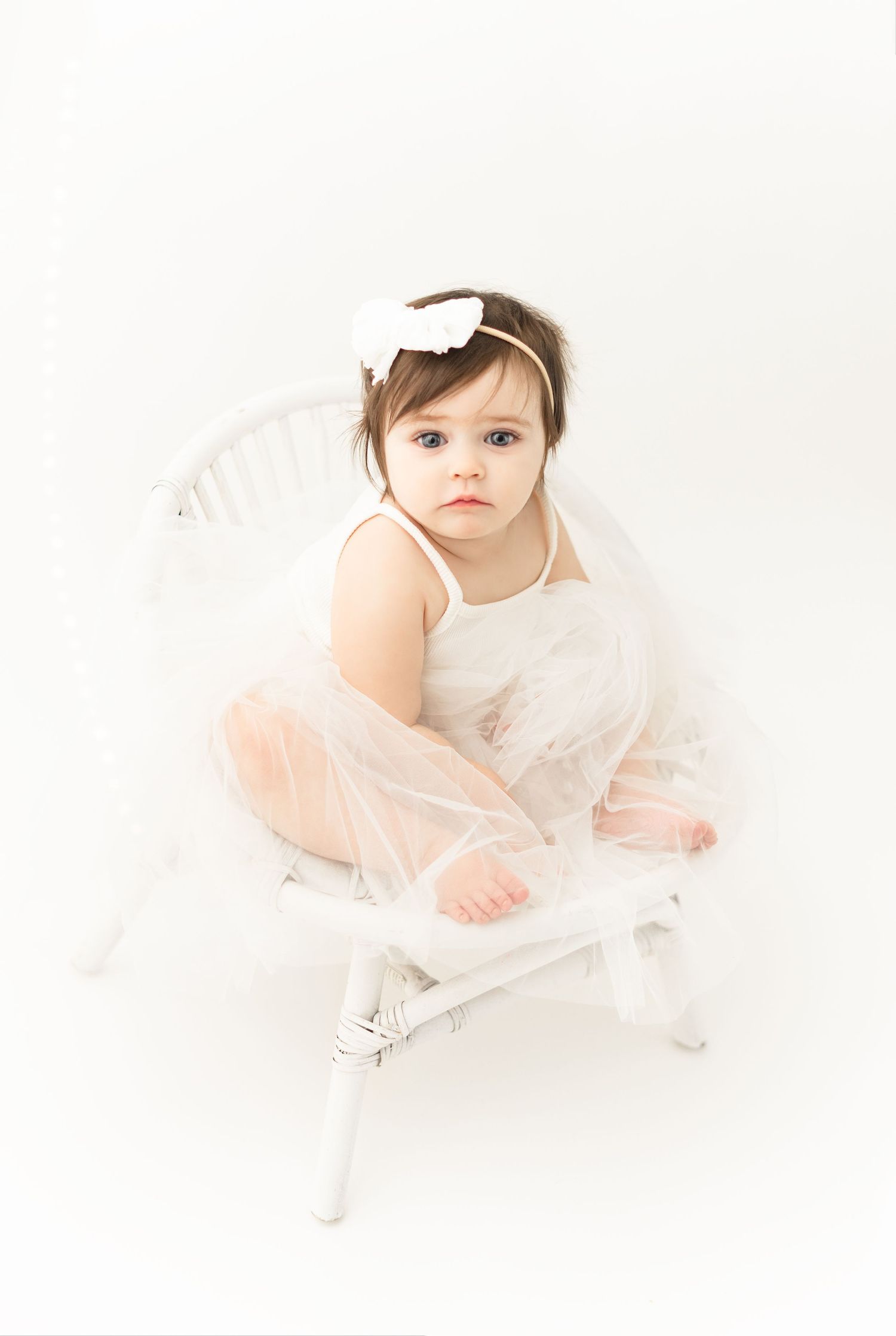 Small child in a white tulle dress and bow sits on a vintage white stool against neutral background.