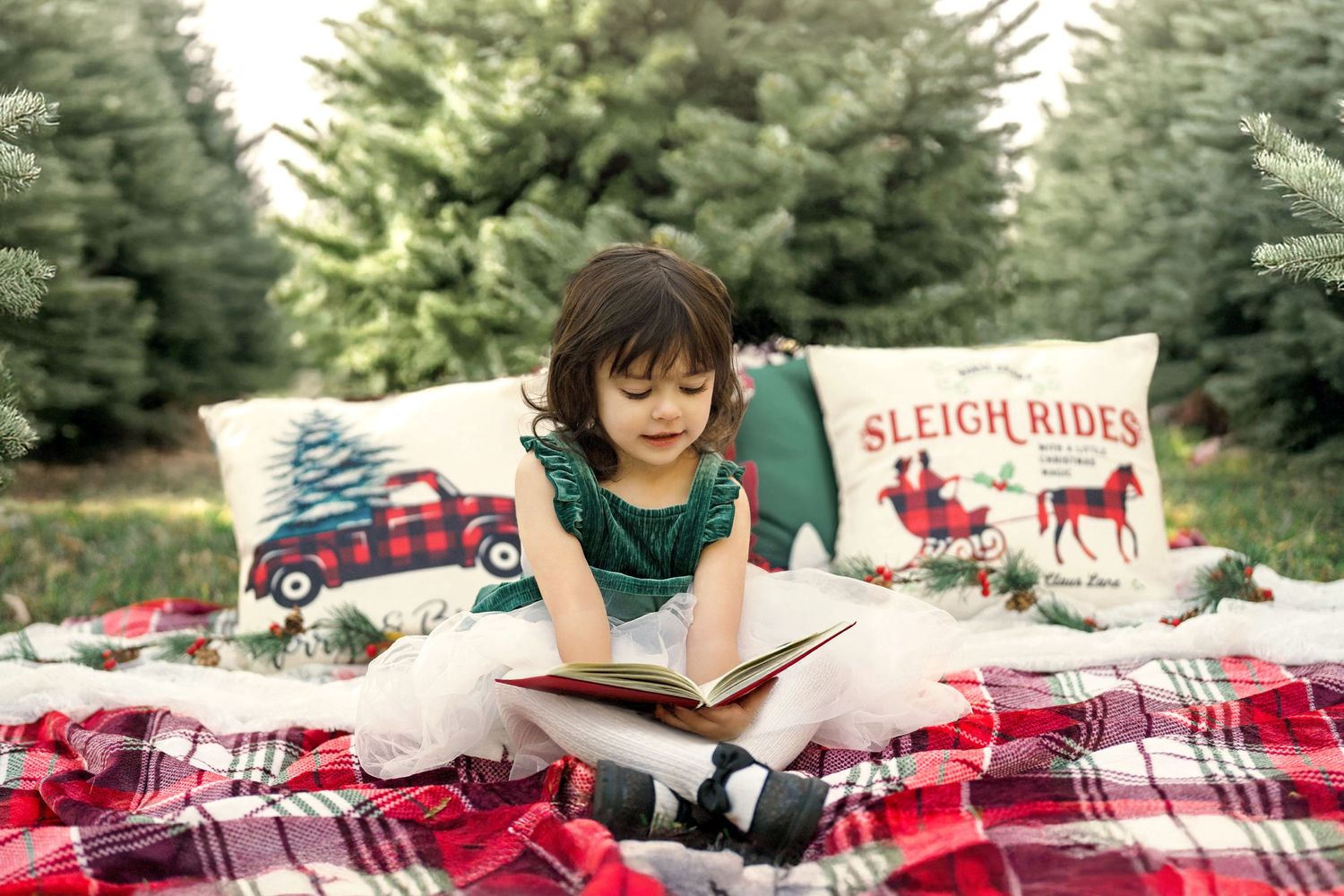 A young girl sits on a festive blanket, reading a Christmas storybook.