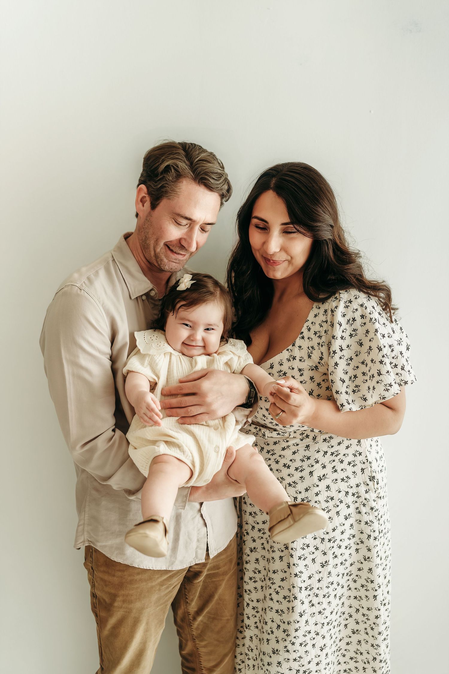 Parents standing together holding their baby girl in a soft, neutral studio photo session.