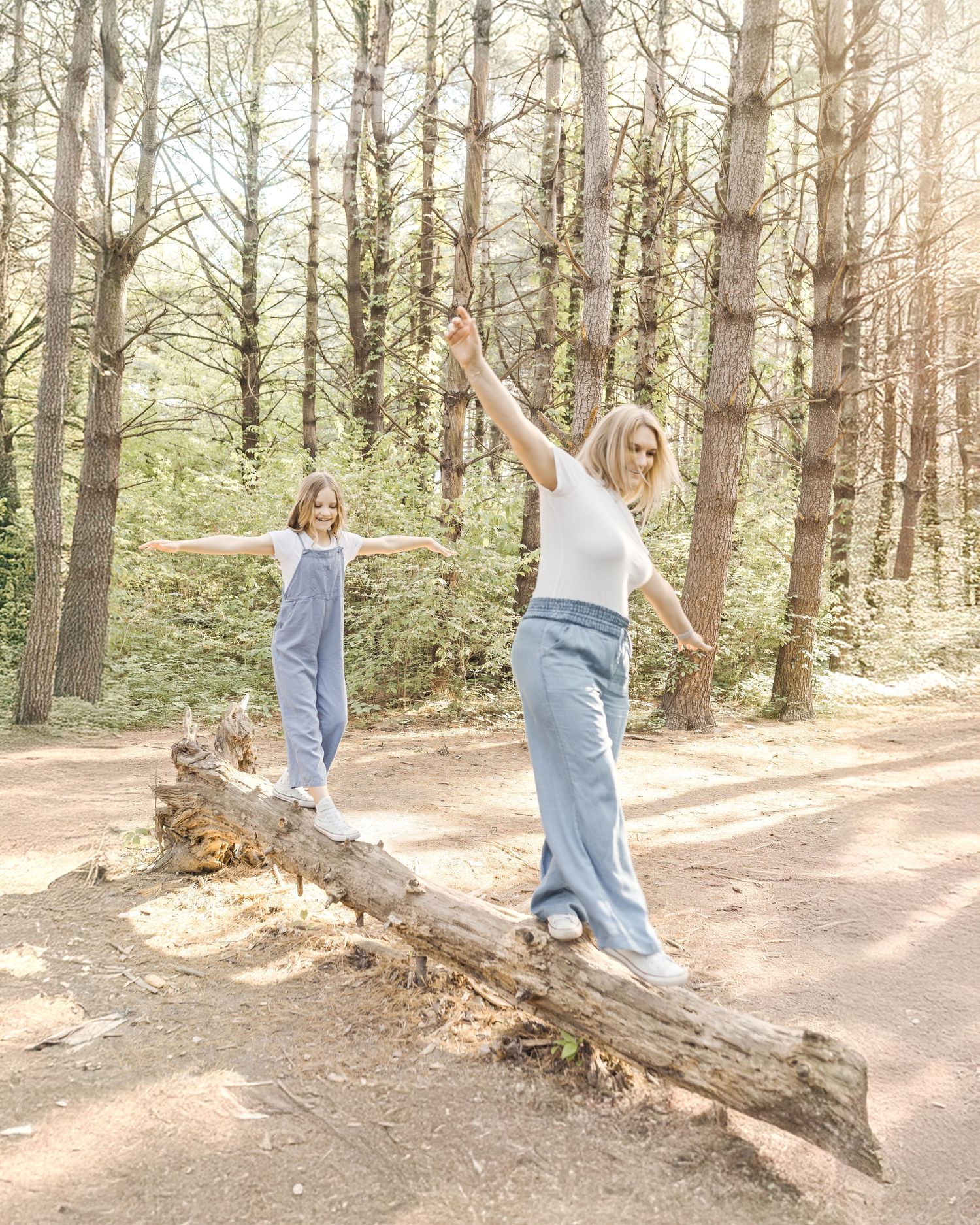 Two people balance on a fallen log along a sunlit forest path while enjoying time outdoors together.