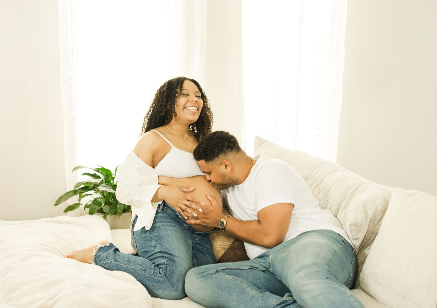 Tender maternity moment with couple wearing blue jeans sitting together on white couch near indoor plant.