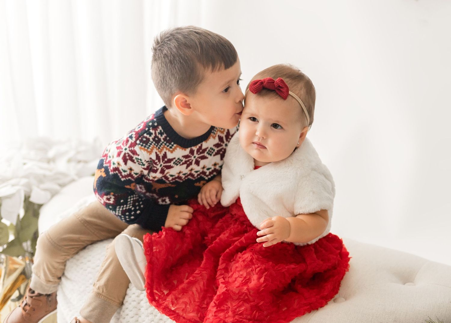 Two children in holiday attire sit together on light backdrop, with one wearing a patterned sweater and other in red dress.