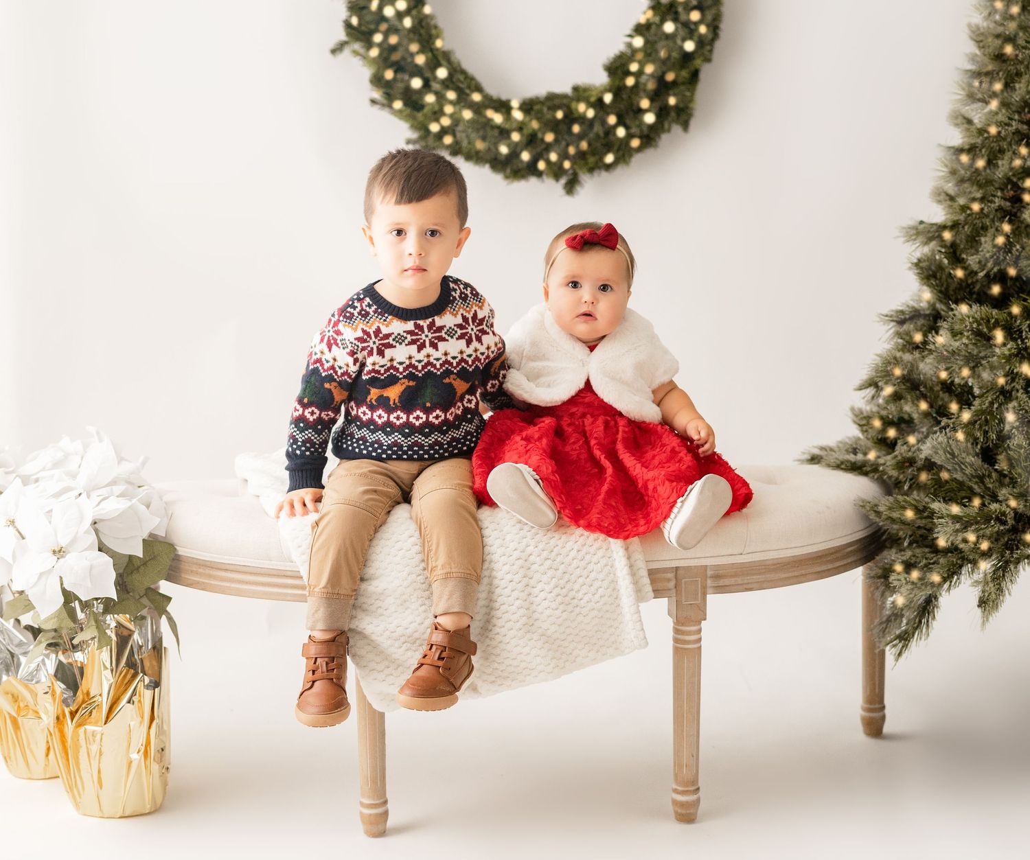 Two small children sit on a bench for Christmas photos with a wreath and decorated trees in the background.