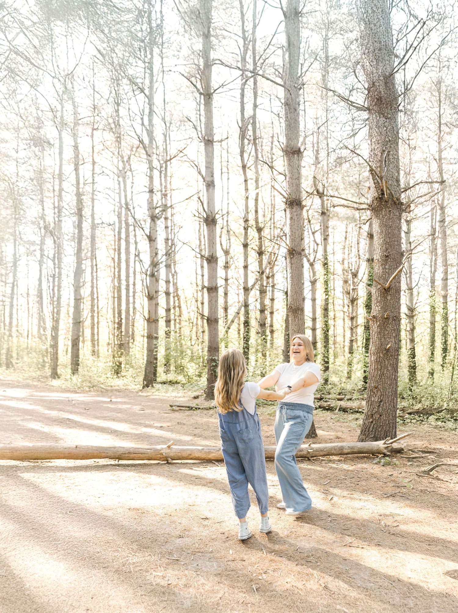 Two people in denim outfits dancing together on a sunlit forest path surrounded by tall pine trees.