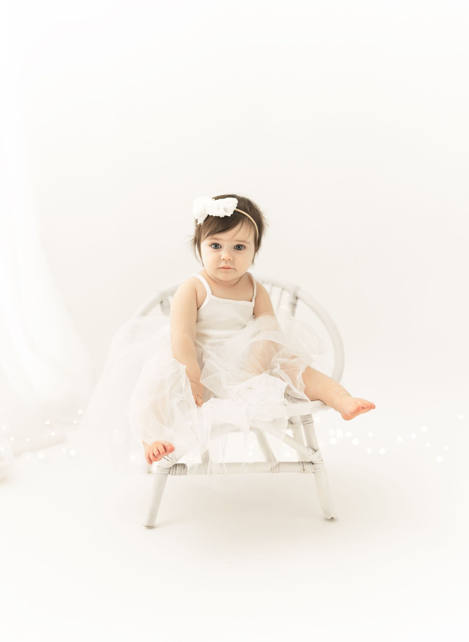Baby in white dress sits on a small white chair against neutral background in minimalist portrait setting.