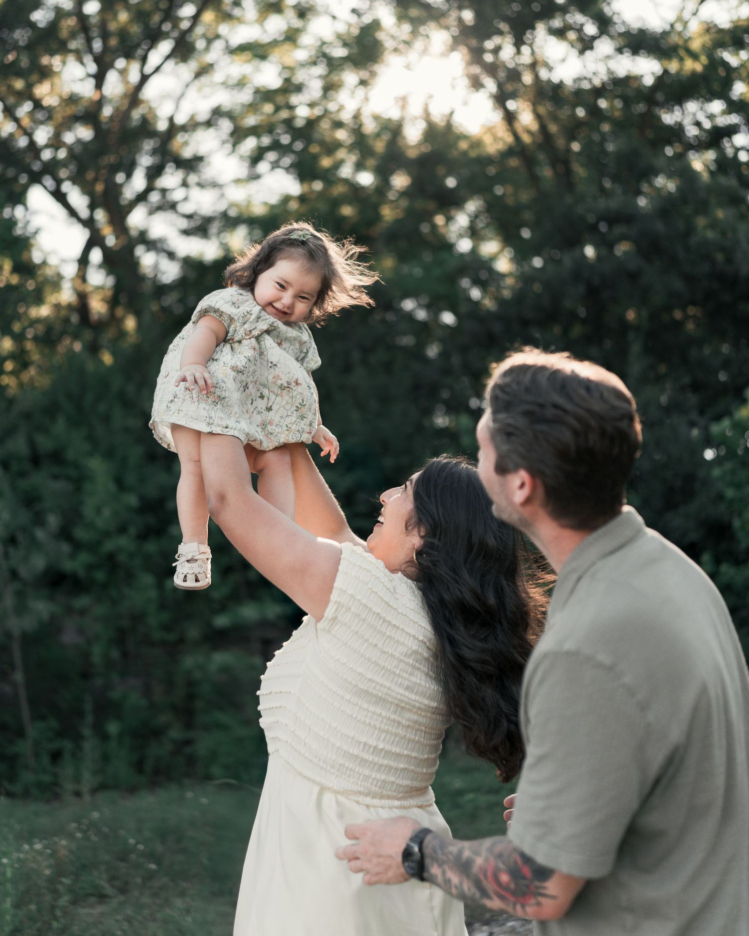 Father lifting his smiling toddler into the air while mom watches during an outdoor family session.
