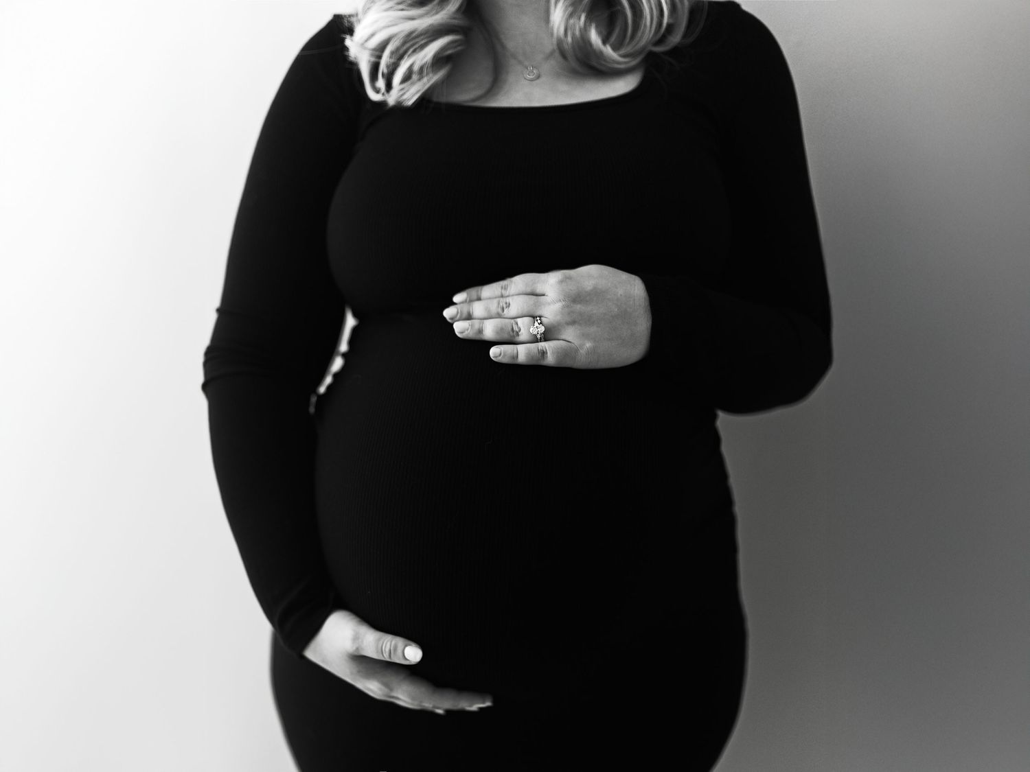 Black and white maternity photo of pregnant woman in black dress cradling baby bump with ringed hands.