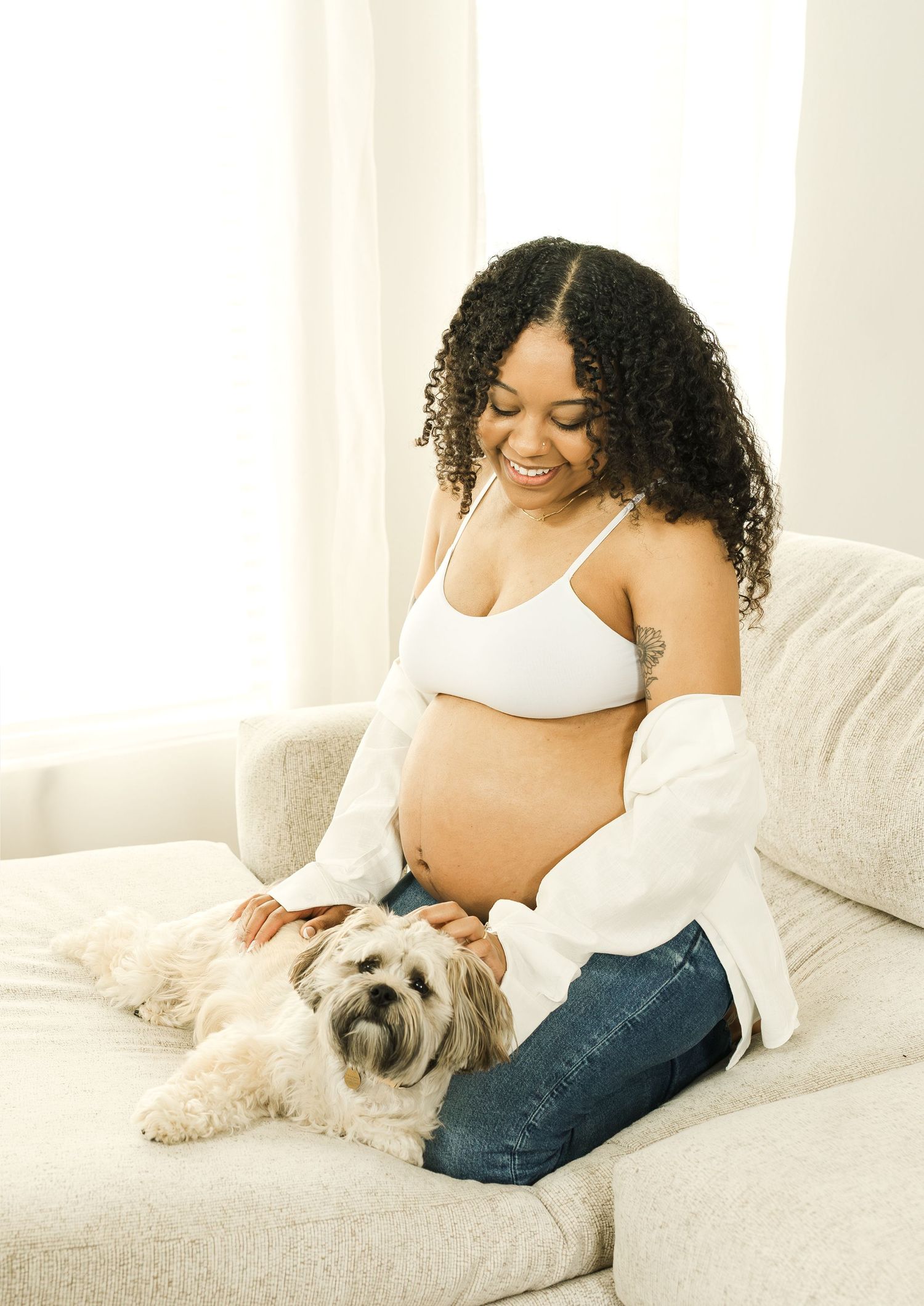 Pregnant woman in white top and jeans lounges on beige couch with small white fluffy dog in peaceful maternity photos.