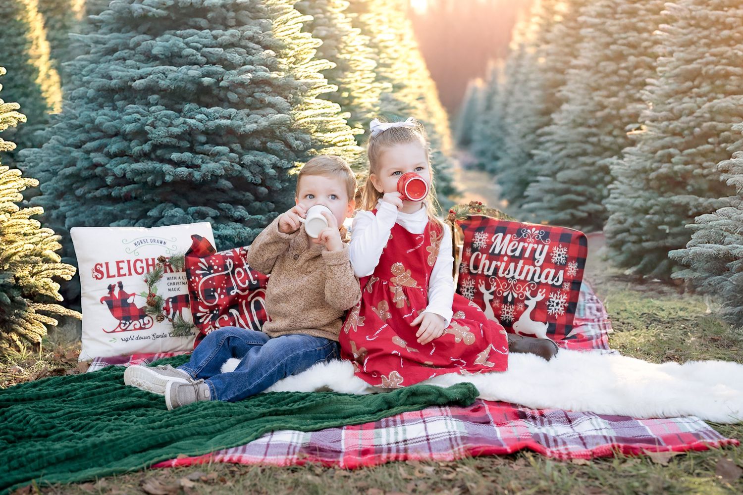 A young boy and girl sit on a festive blanket among Christmas trees, each holding a cup of hot cocoa.