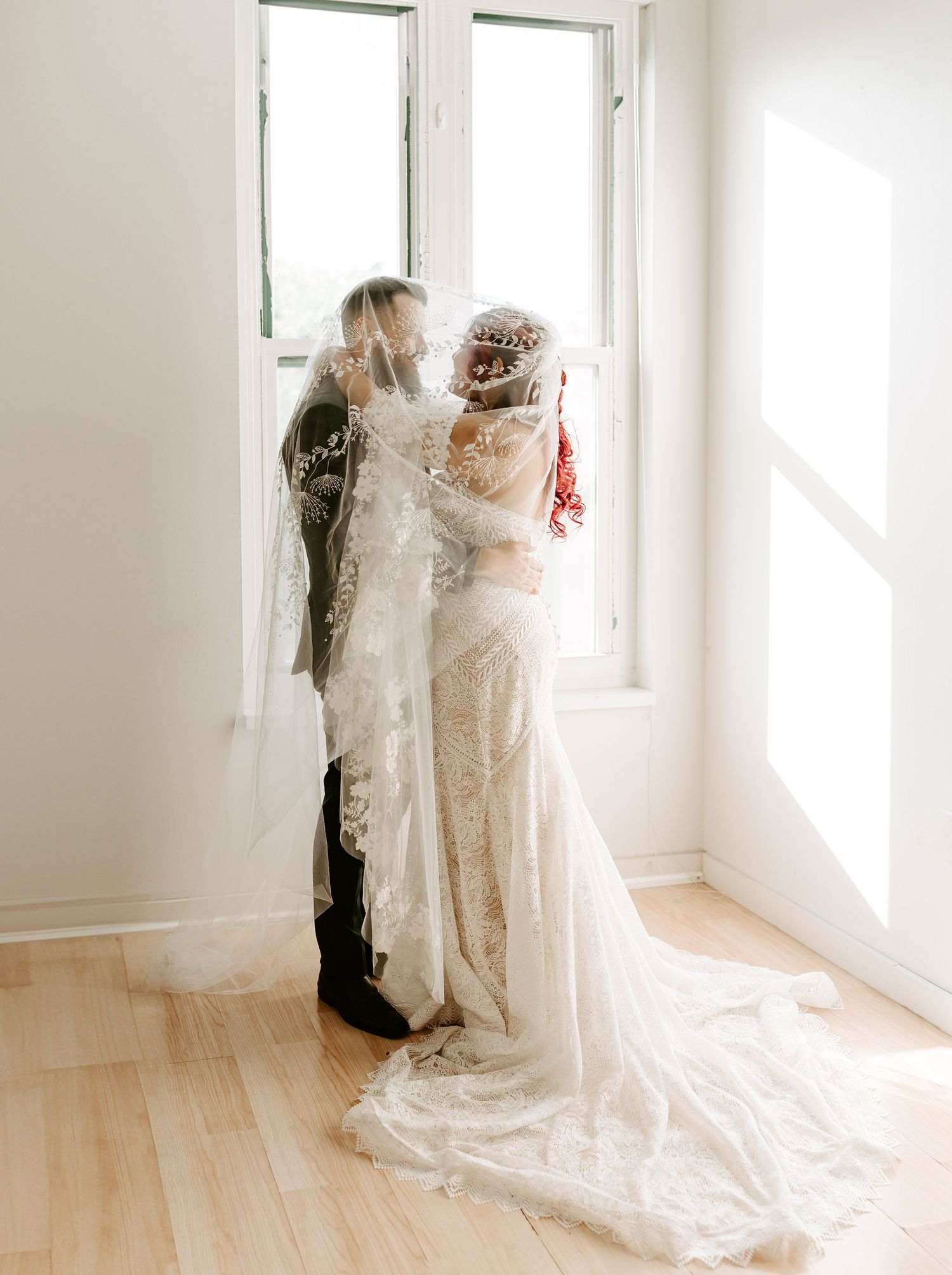 A couple in wedding attire stands together by bright windows in a minimalist white room with hardwood floors.