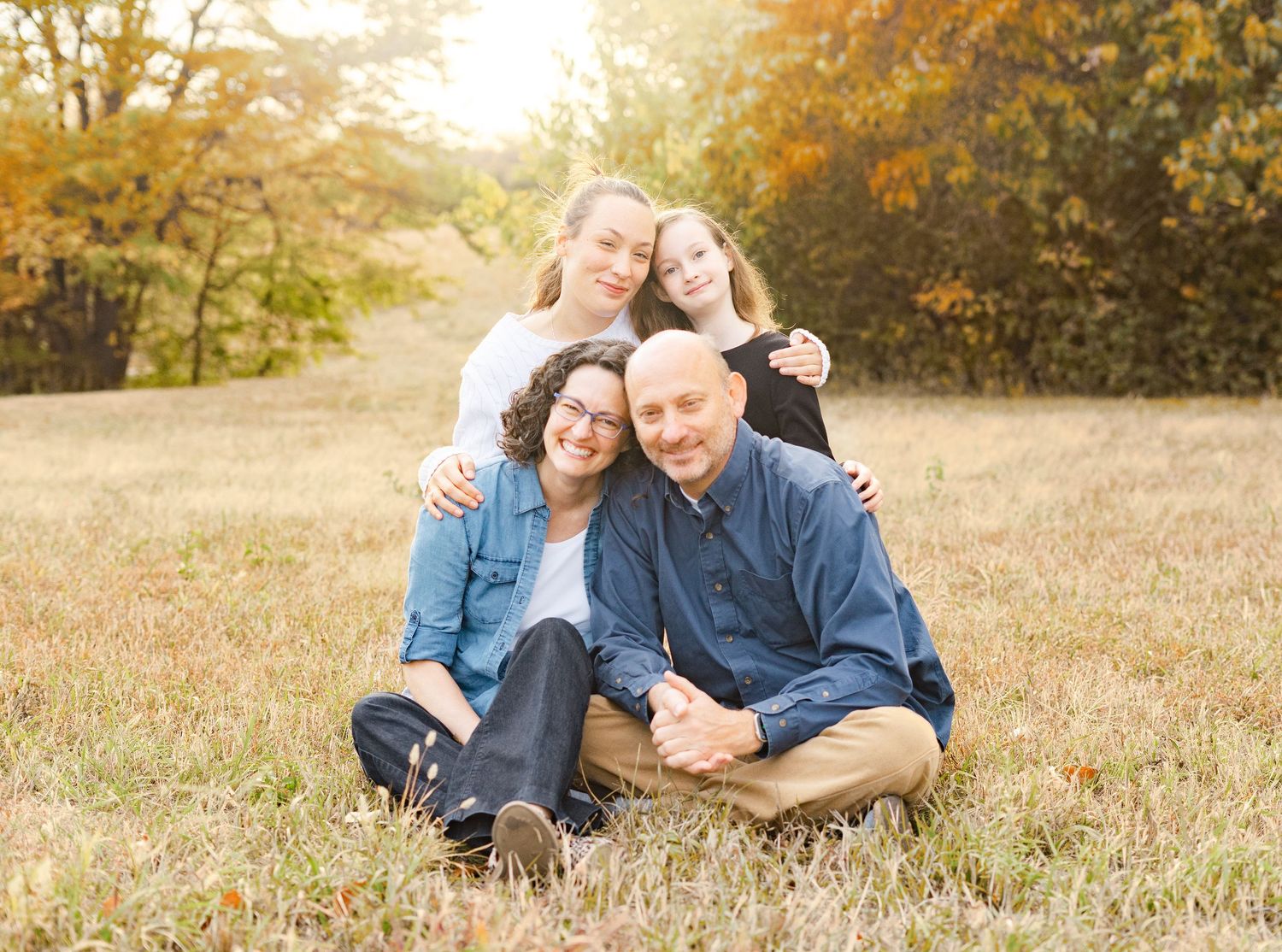 Family of four sits together in autumn field with golden sunlight streaming through trees in background.