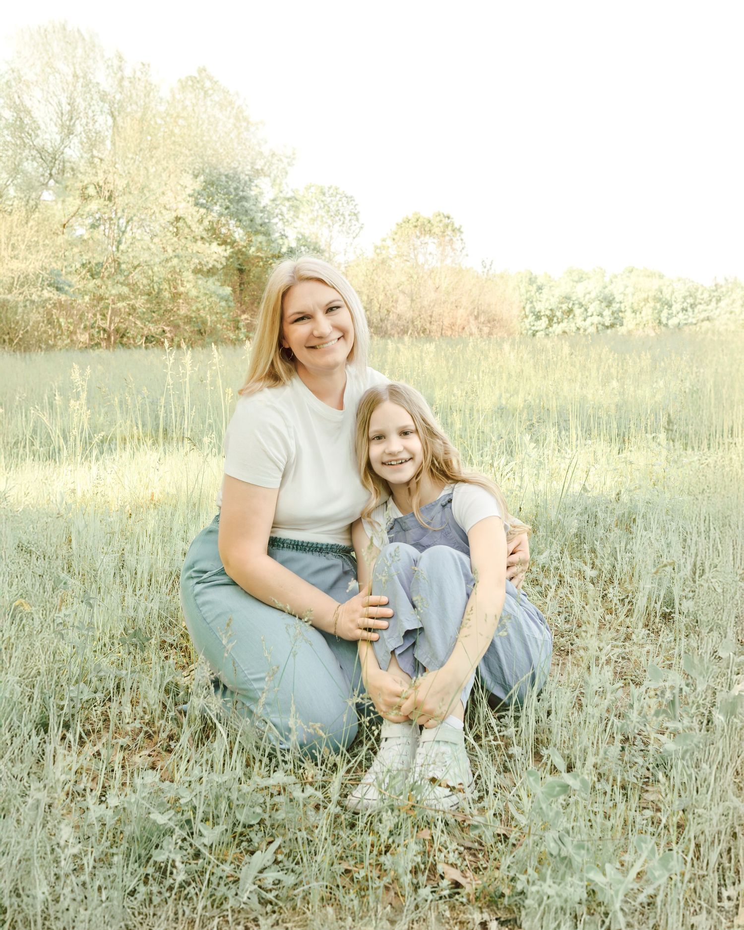 Two people in jeans sit together in a sunlit field of tall grass during golden hour.