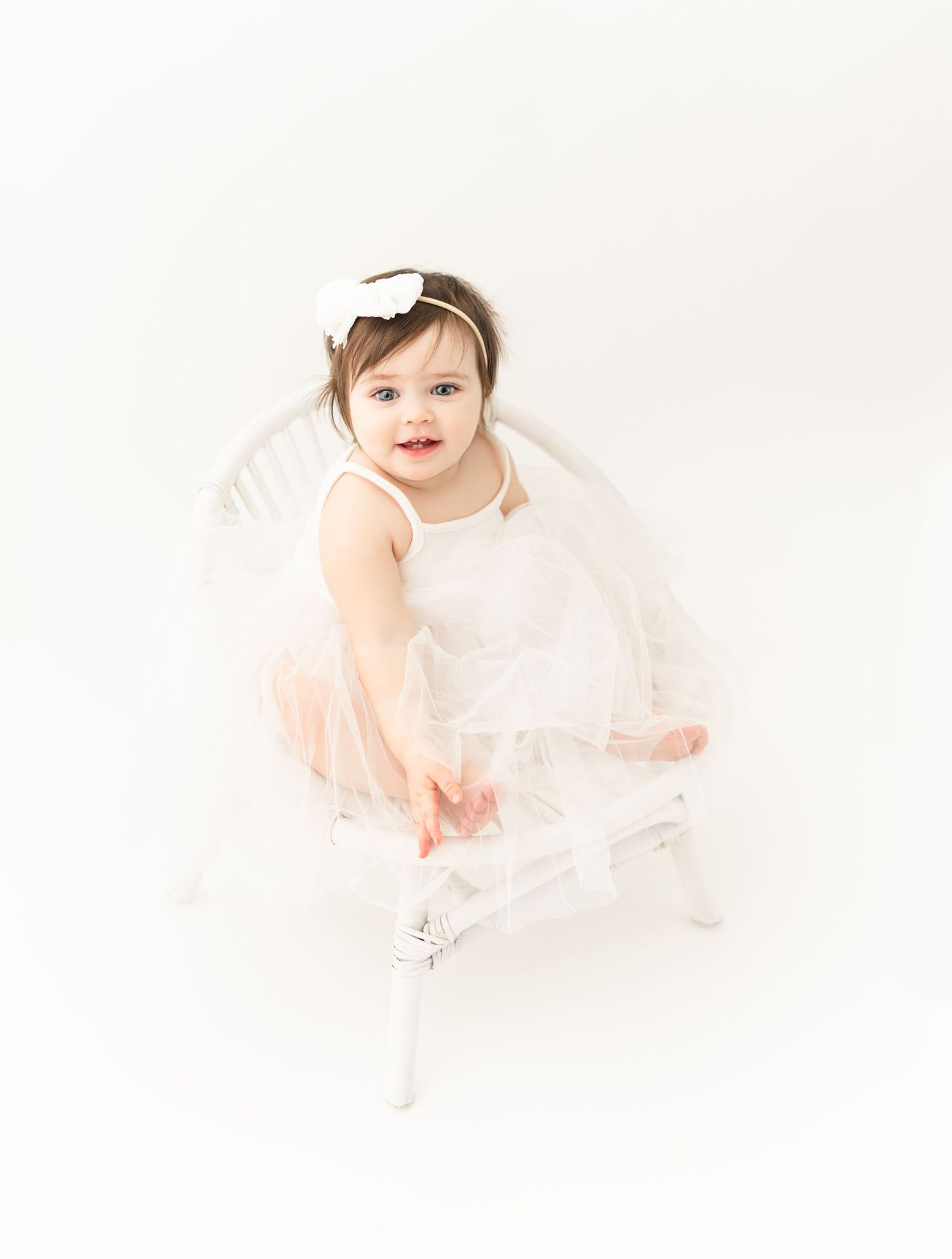 Sweet baby wearing a white tulle dress and bow sits on white backdrop during portrait session.