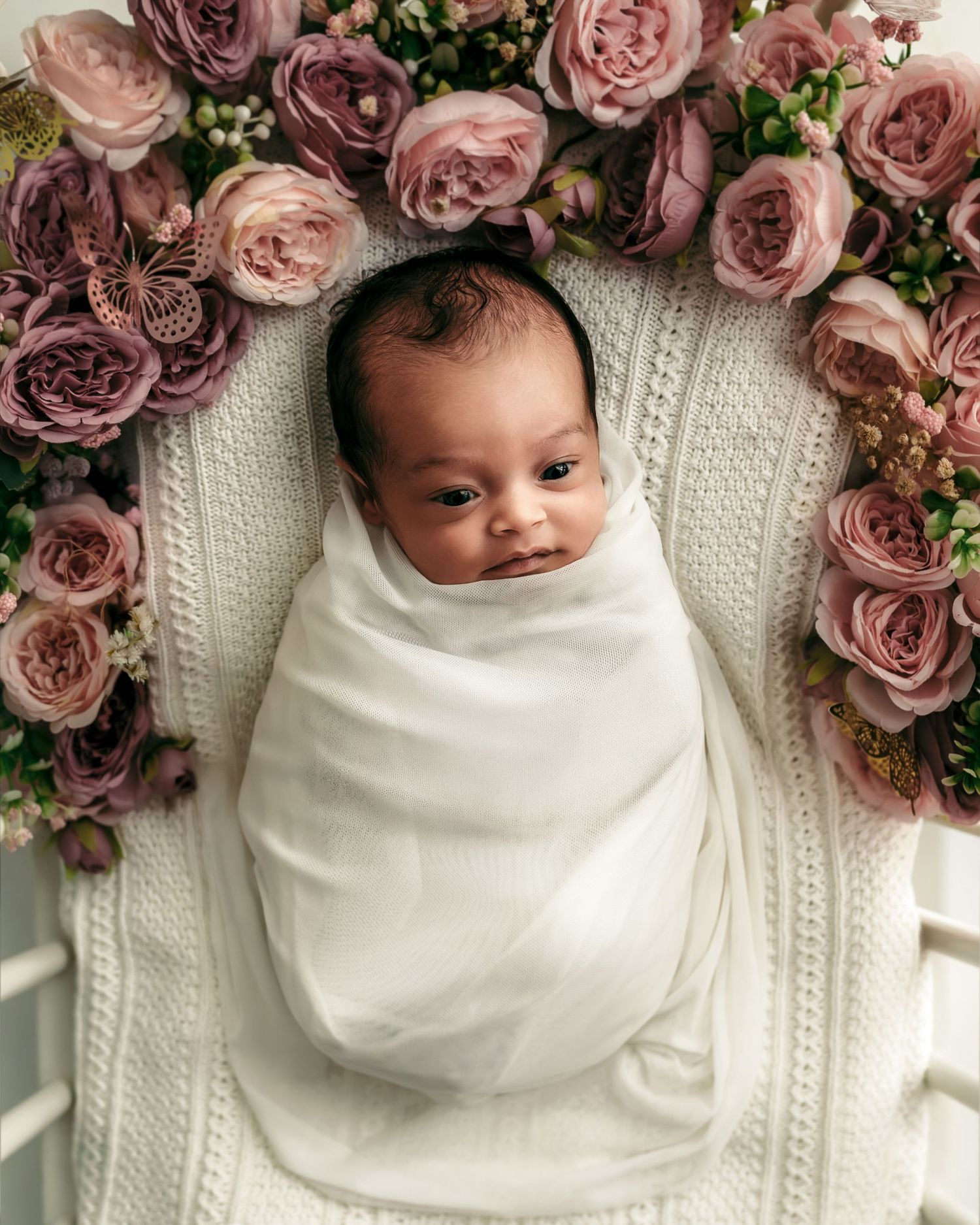 Newborn baby swaddled in a white wrap surrounded by pink and mauve flowers during a studio newborn photography session in Kansas City.