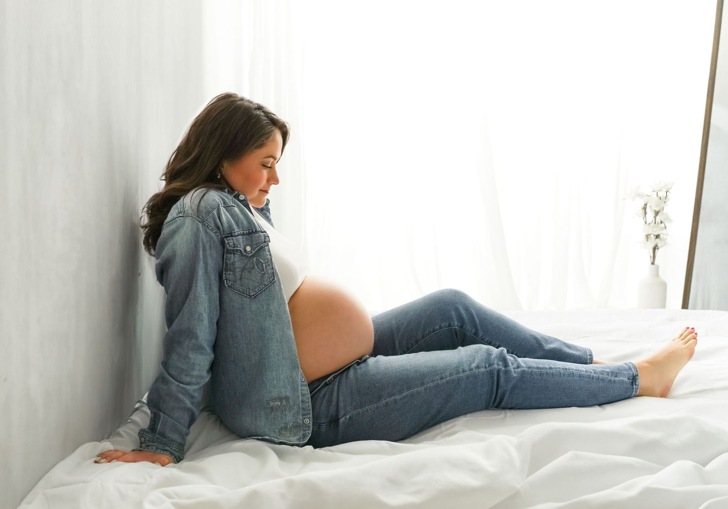 A pregnant woman relaxes on white bedding wearing denim shirt and jeans while leaning against a wall.