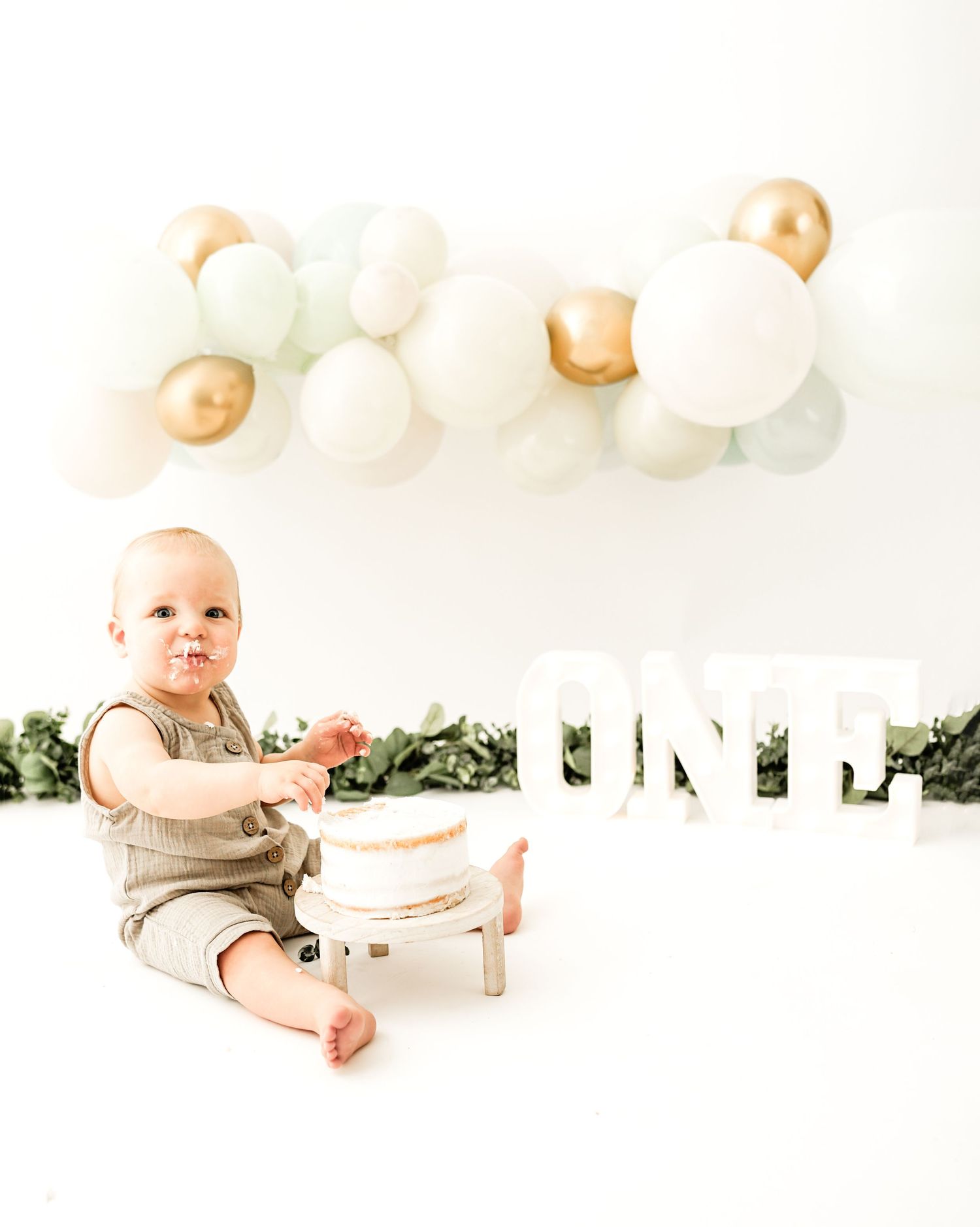 First birthday celebration setup with neutral balloons, greenery, and white minimalist decor on white background.
