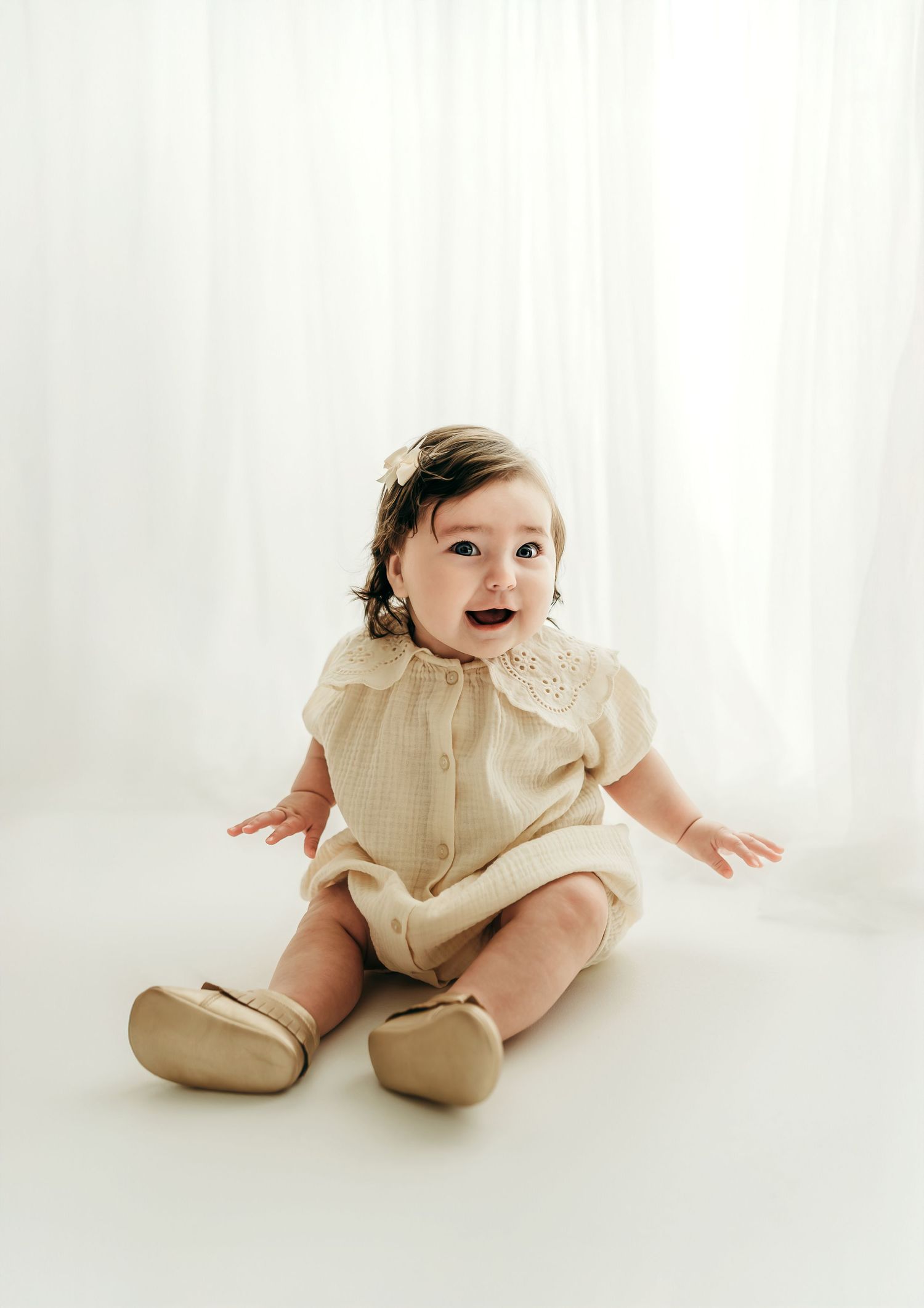 A baby girl around 6 months old sitting and smiling on a white backdrop, wearing a cream outfit in a soft, minimal studio milestone session.