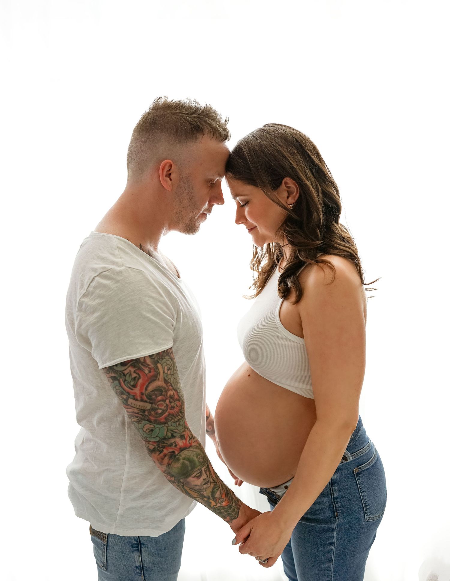 A pregnant couple in white tops and jeans stand together lovingly looking down at the baby bump in multiple poses.