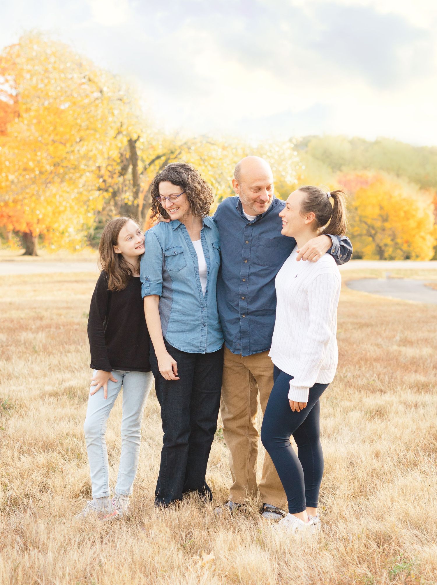 A group of four people laugh together while standing in a golden field during autumn with yellow trees in background.
