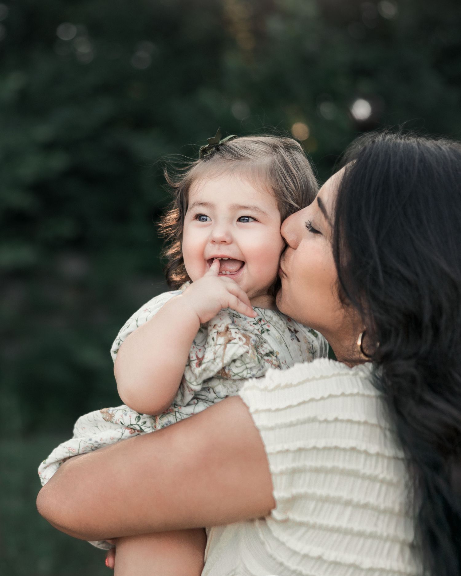 Mother kissing her smiling daughter during a candid outdoor family portrait.