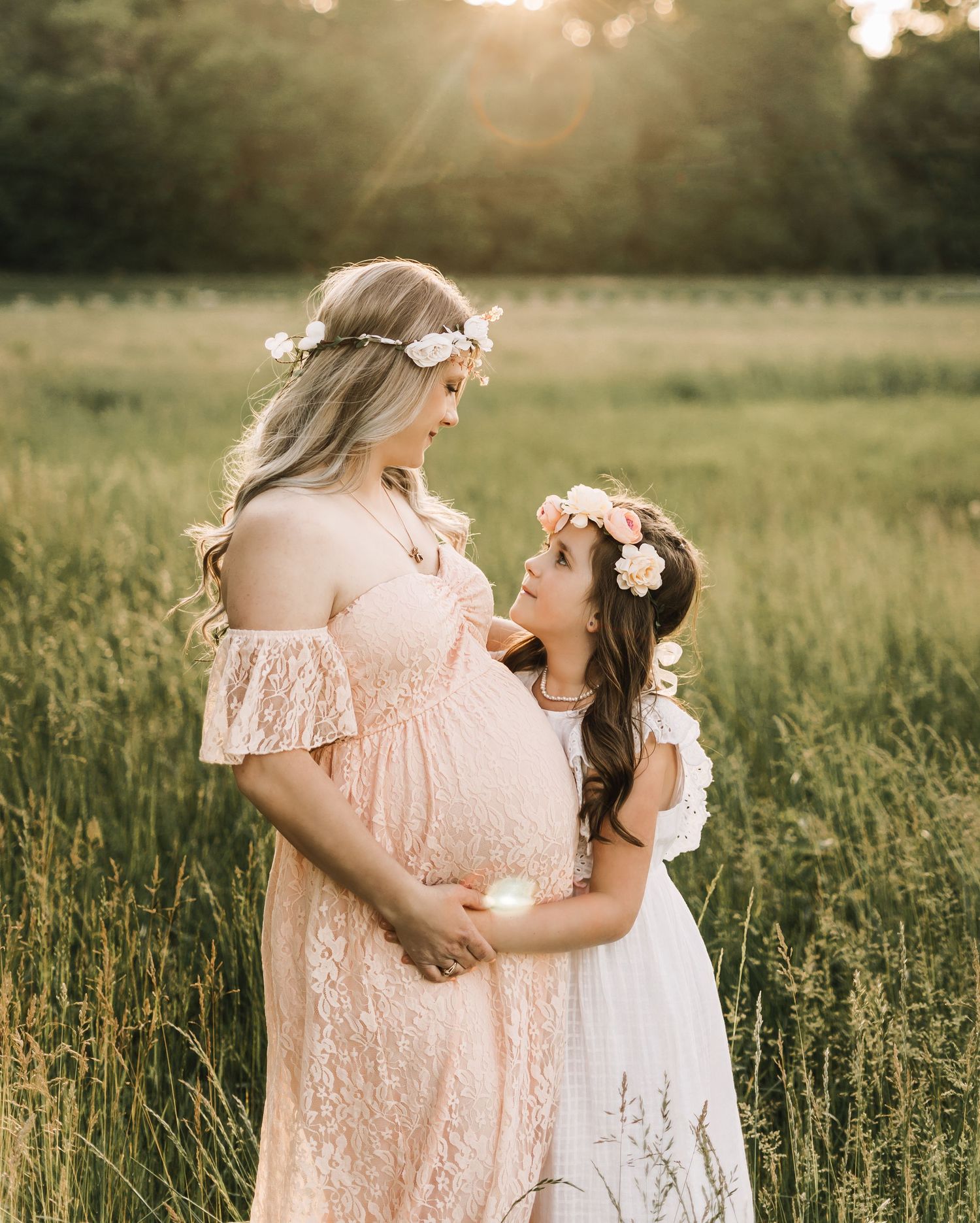 A pregnant woman in a pink lace dress and a young girl in white wear floral crowns while posing in a sunlit meadow in kansas city