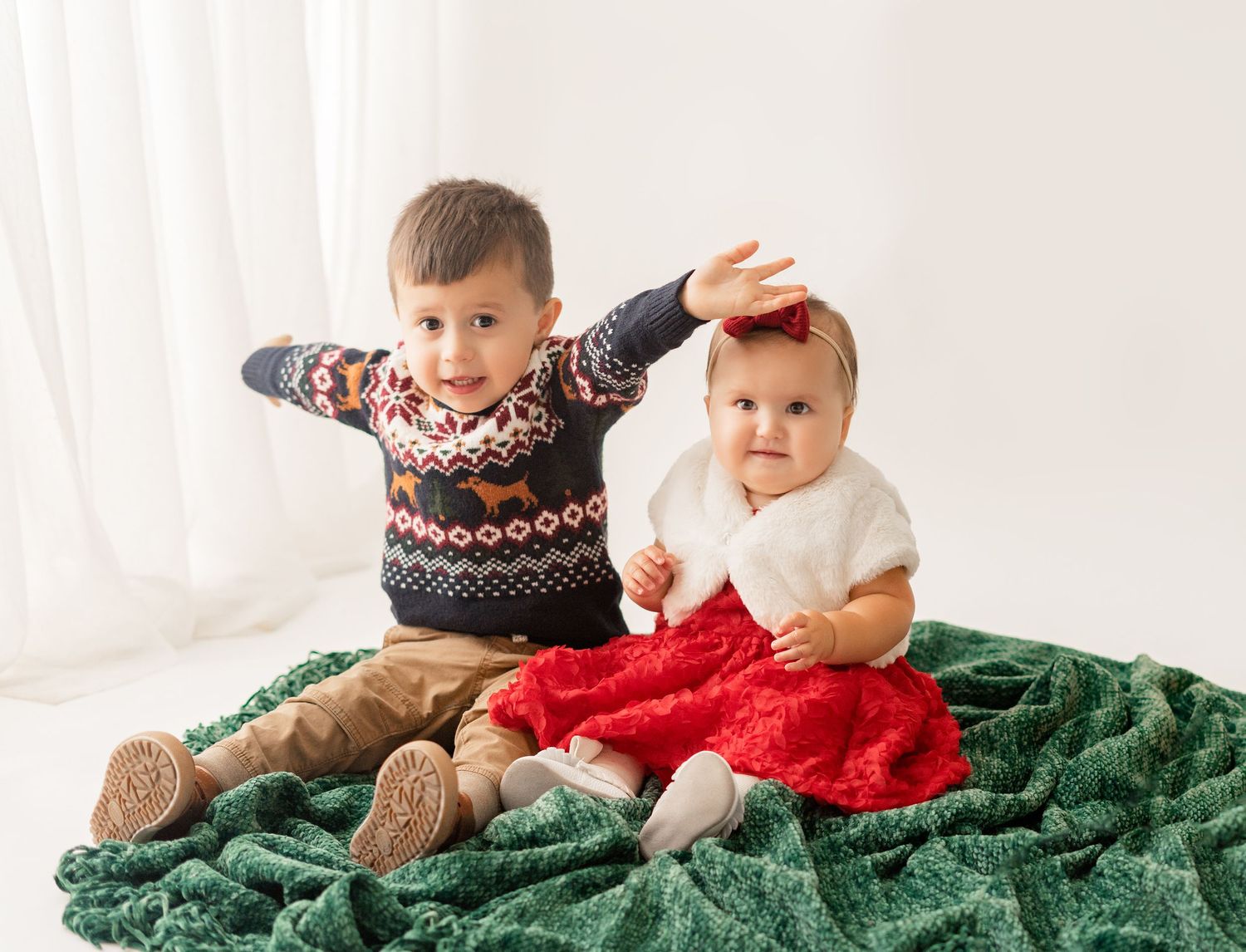 Two children sit on a green blanket wearing winter sweaters during a holiday photo session.