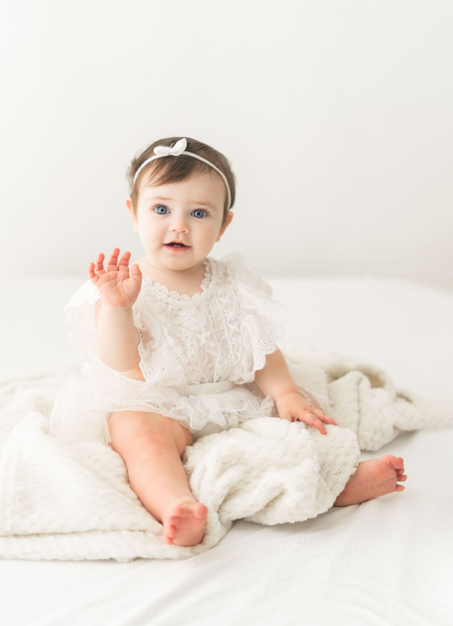 Sweet baby in white lace romper and bow headband sitting on soft white blanket with waving hand.
