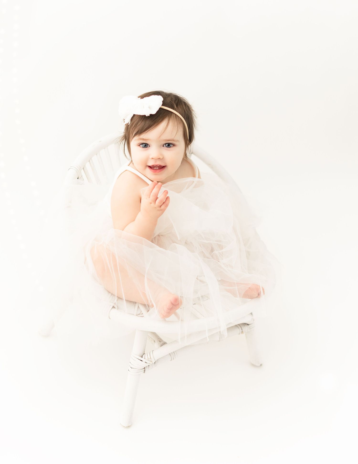 Baby in white tulle dress smiling while sitting in vintage chair against white background.