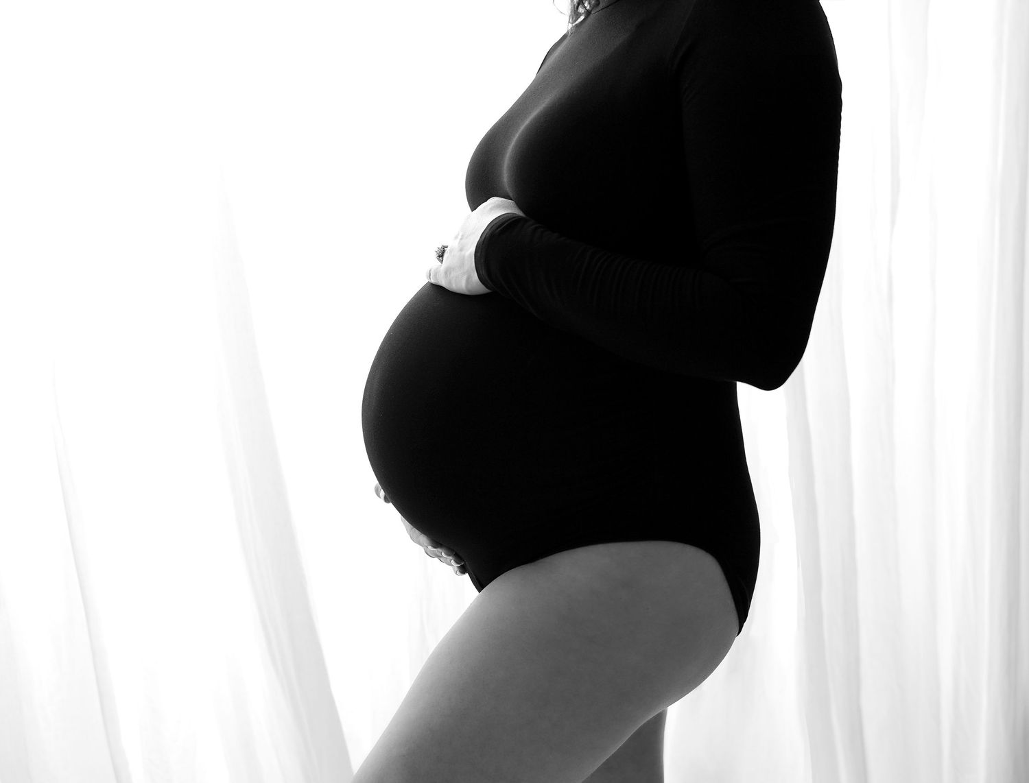 Black and white maternity photo of pregnant woman in bodysuit cradling baby bump by sheer curtains.
