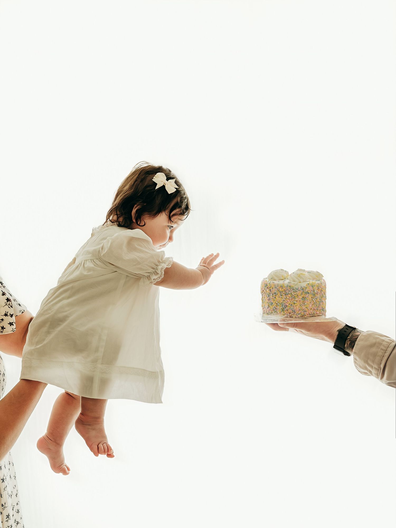 A baby girl reaching toward a small cake while being held by her parents during a soft, neutral milestone photo session.