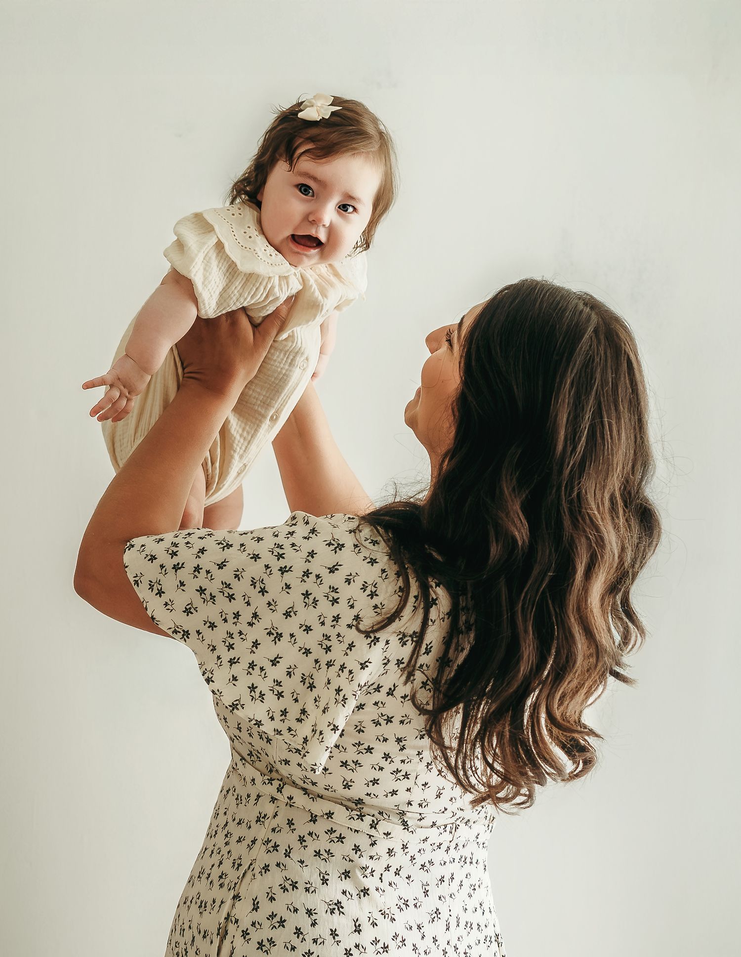A mother lifting her baby girl into the air during a soft, neutral studio photo session.